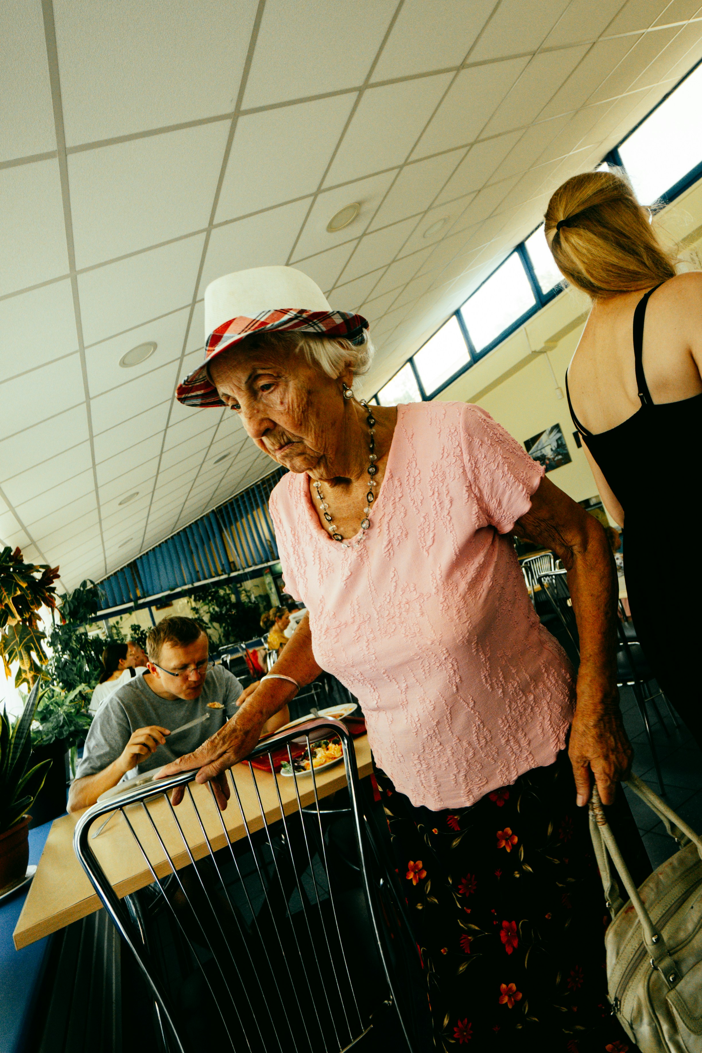 Elderly woman in hat and pink shirt walks past table.