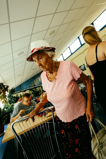 Elderly woman in hat and pink shirt walks past table.