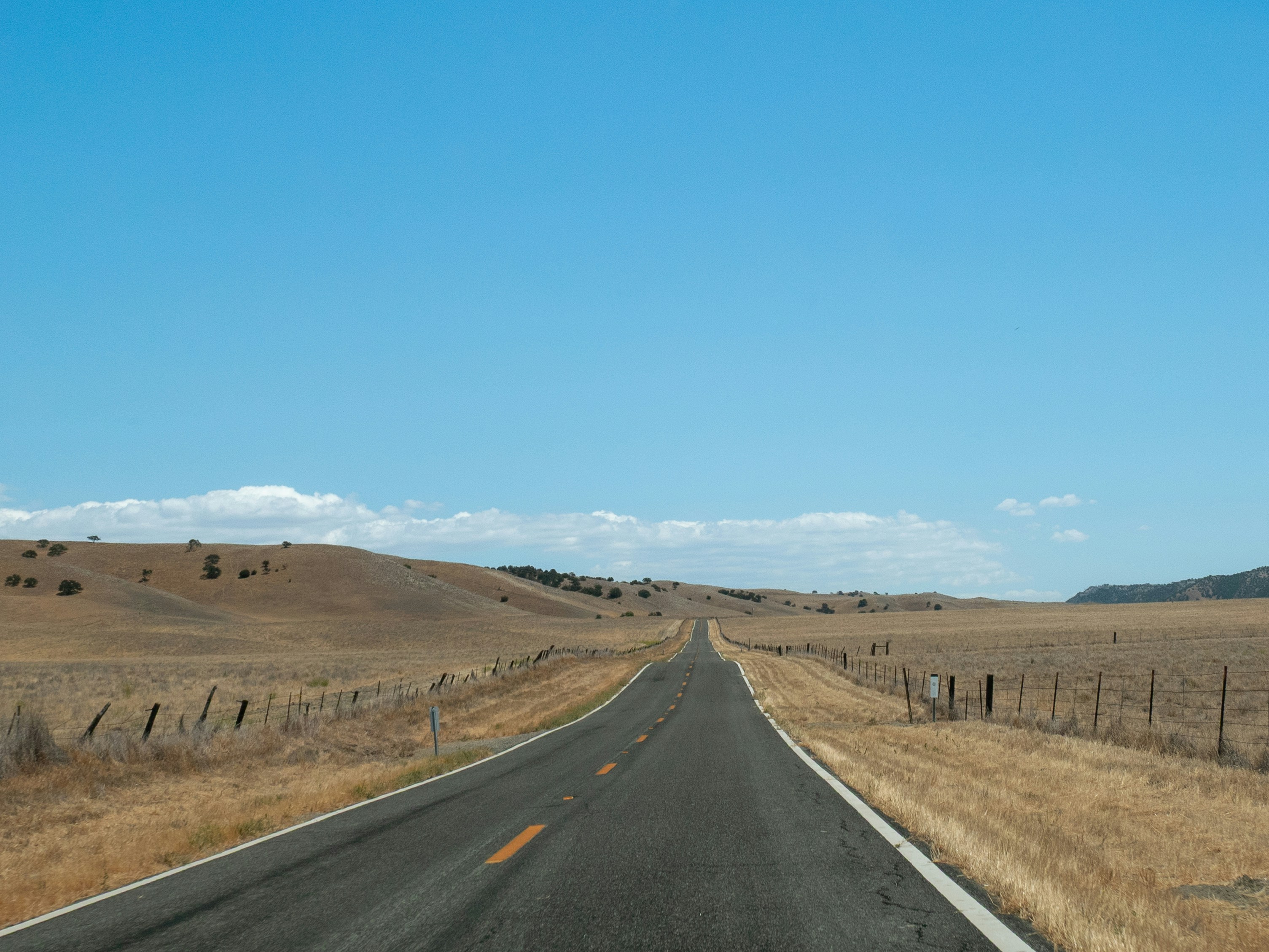 Long empty road through dry grassy hills under blue sky.