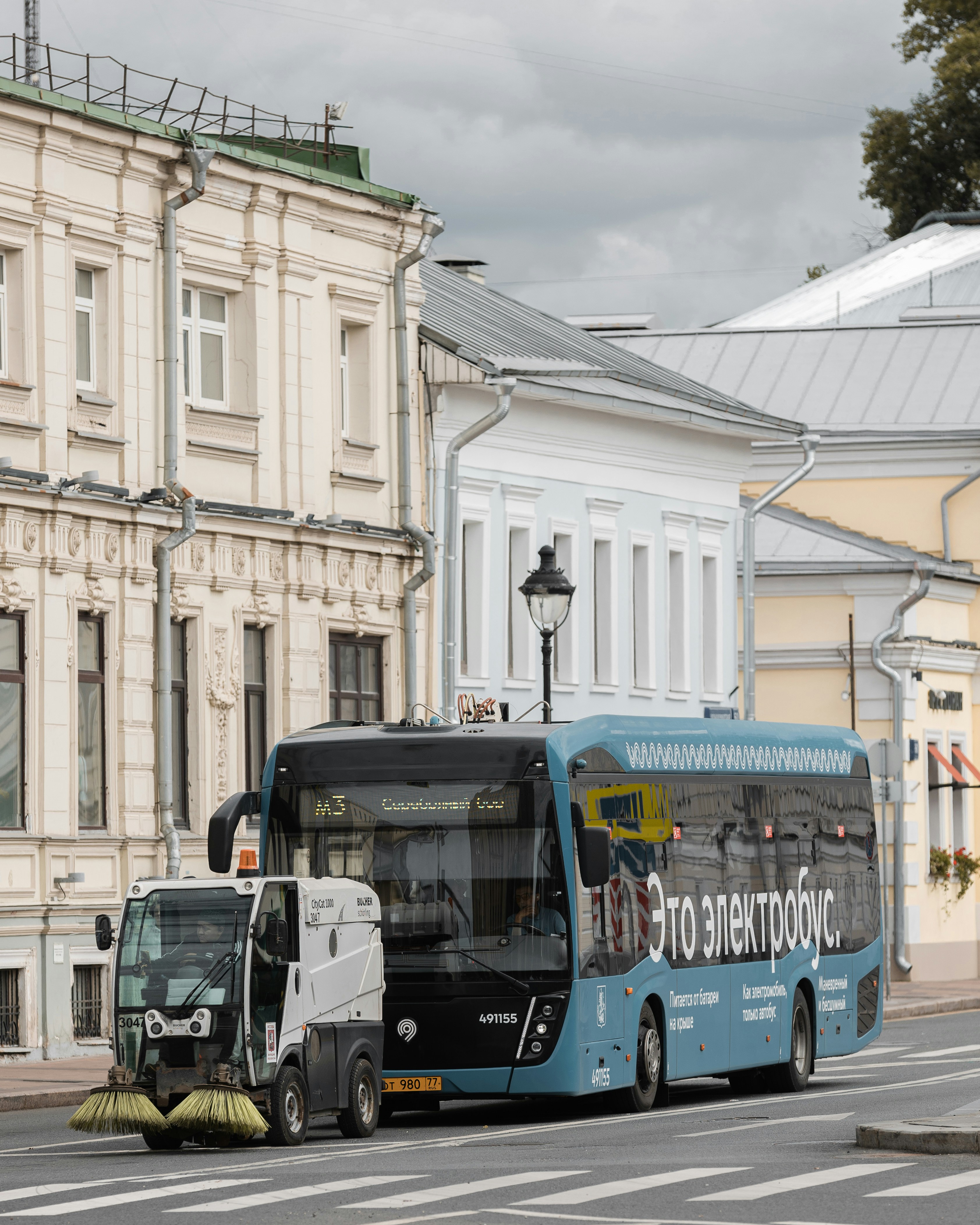 A blue electric bus and street sweeper on a street.