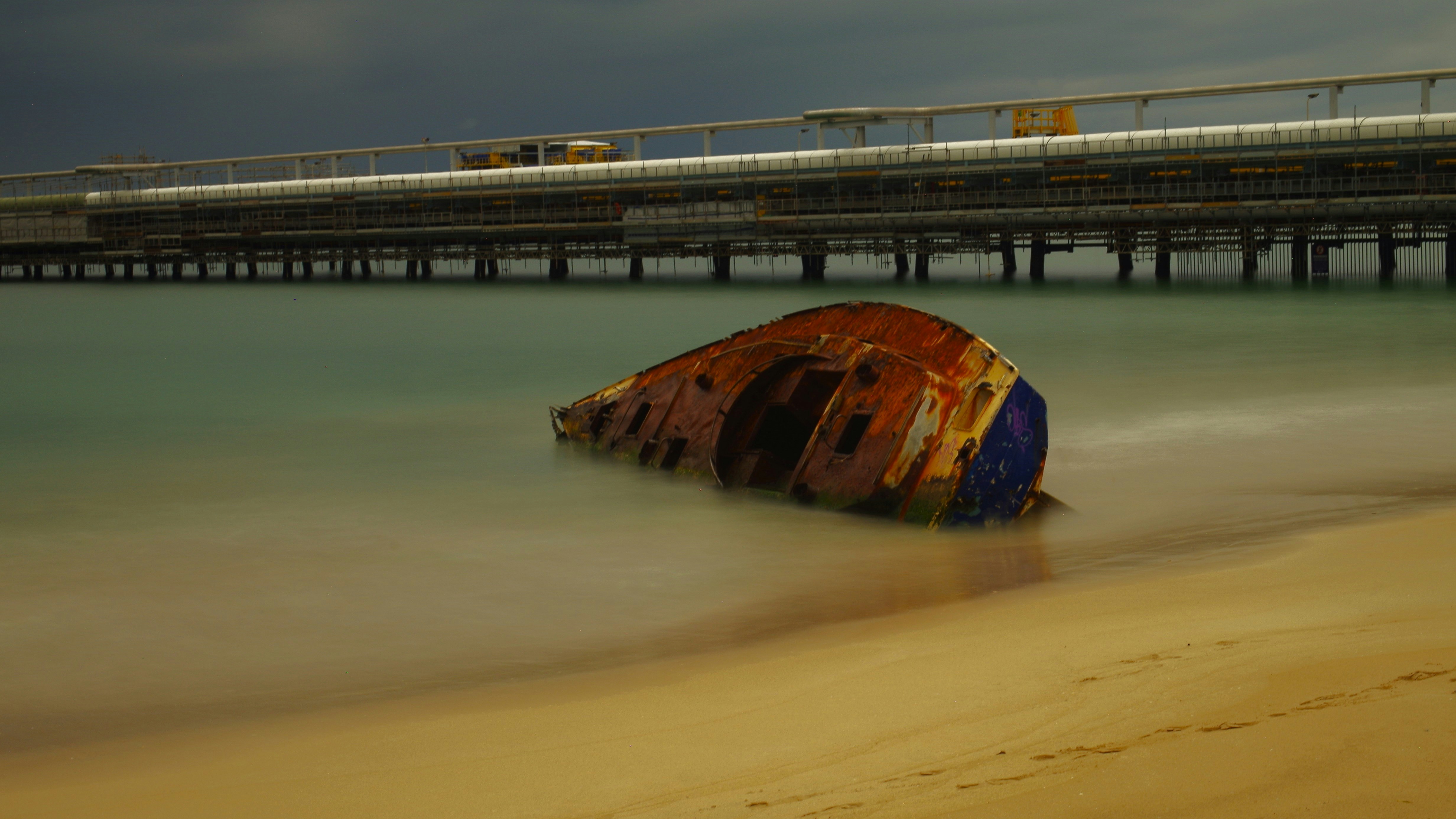 Rusty shipwreck on a sandy beach with pier.