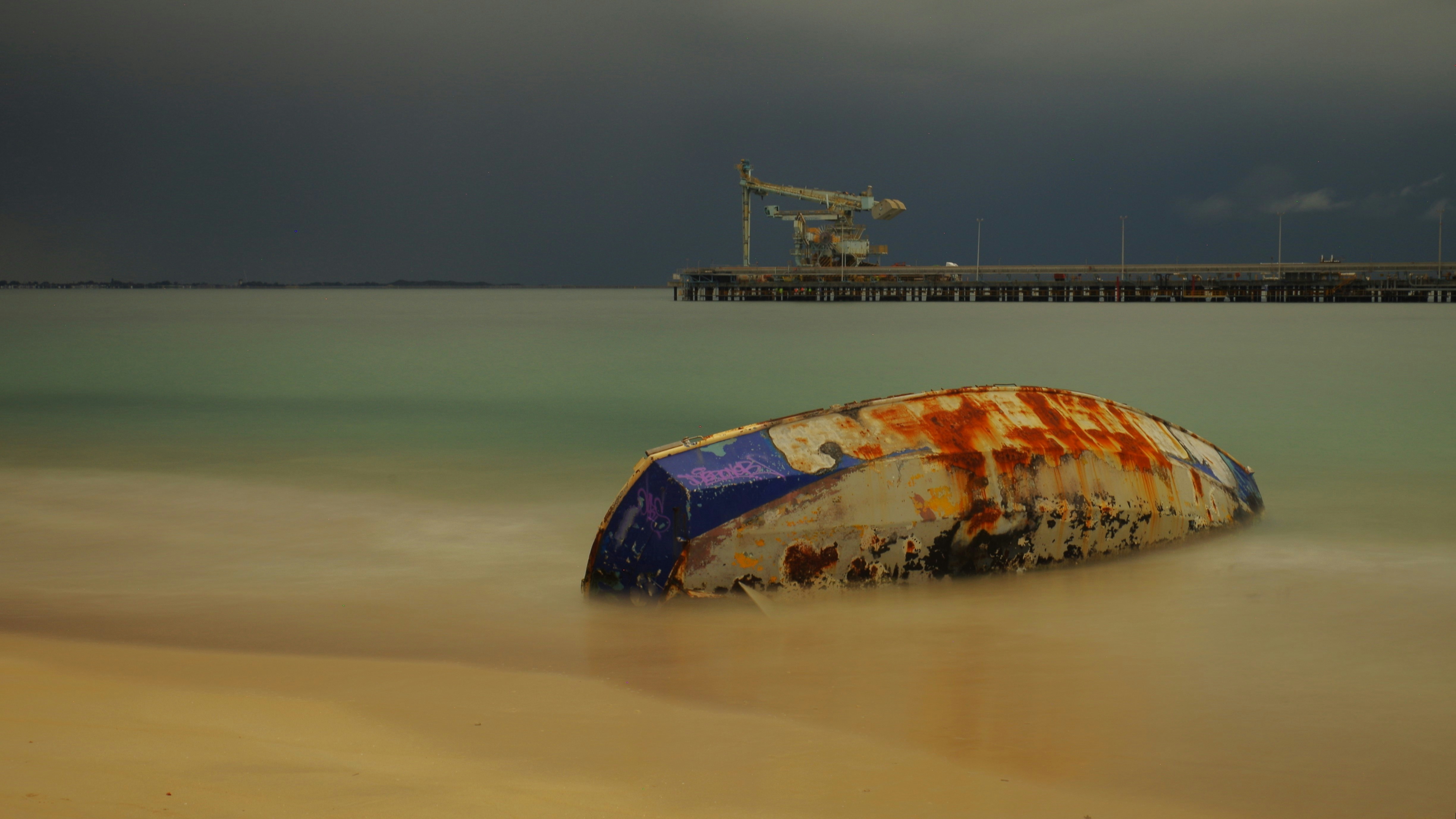 Rusty boat hull washed ashore near a pier.