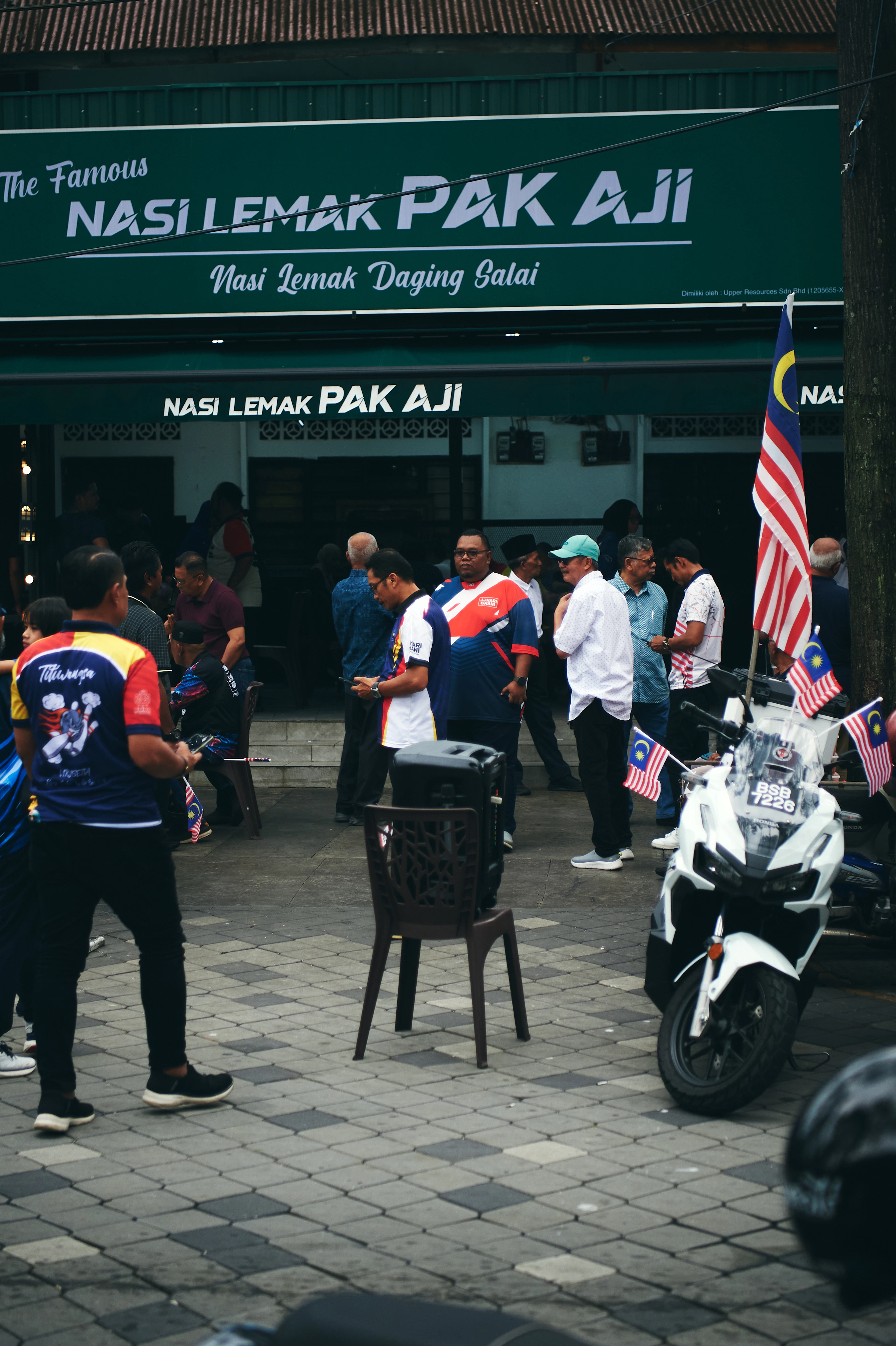 Customers standing and eating on the forecourt of the Nasi Lemak Pak Aji shop in Kampung Baru, Kuala Lumpur, its green signboard overhead and Malaysian flags flying from a motorcycle at the kerb.