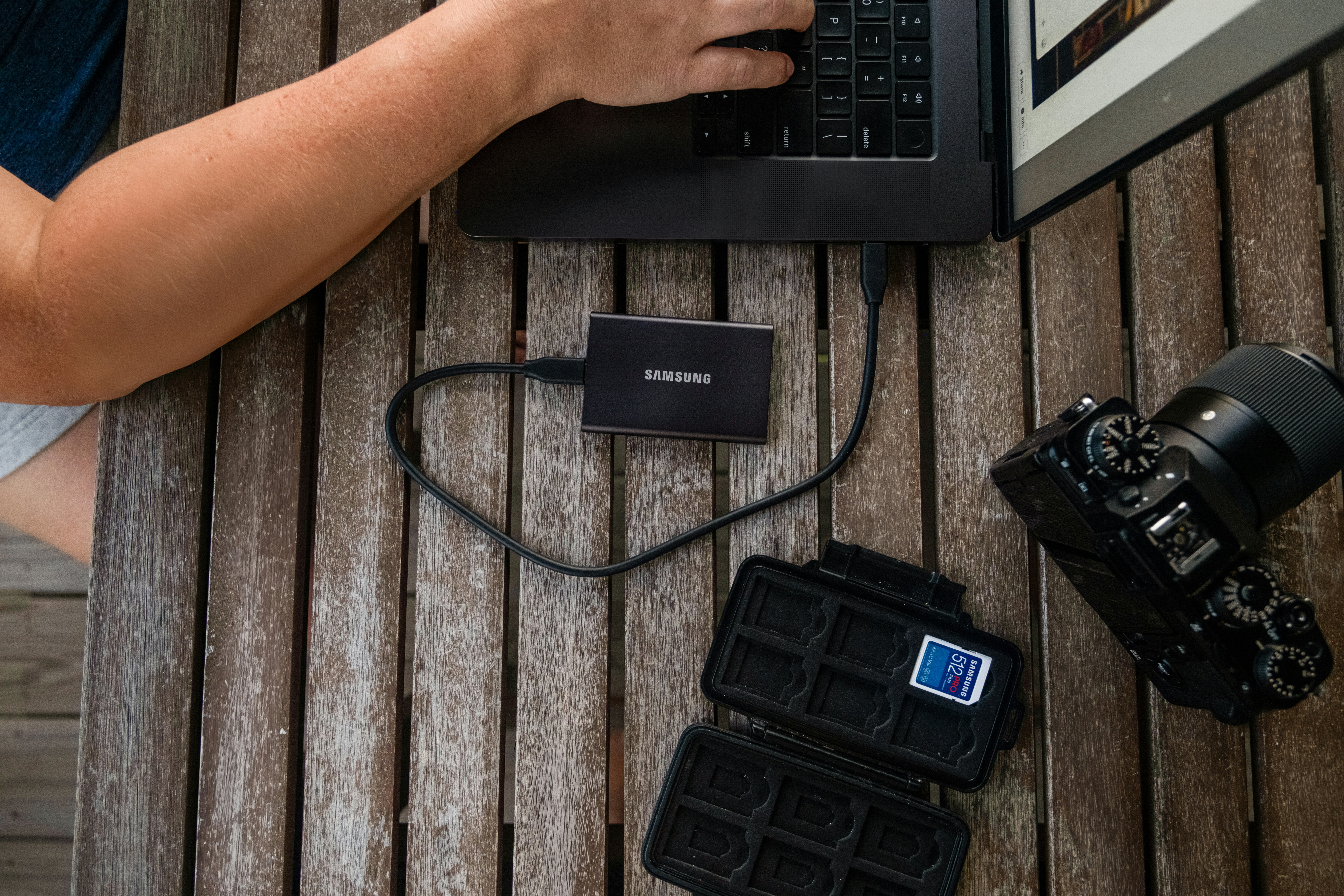 Person typing on laptop with external hard drive and camera.