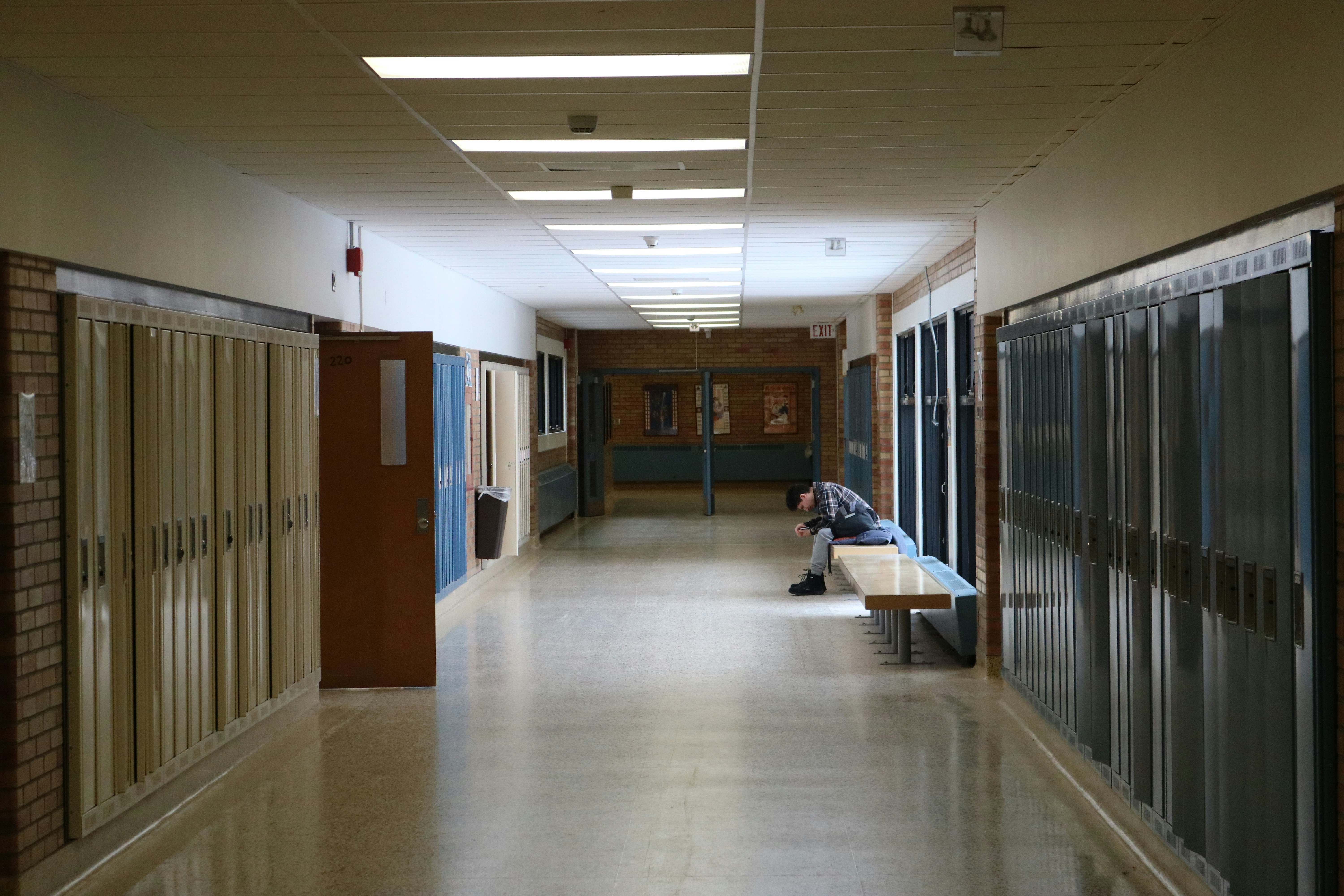 A person sits alone on a bench in a school hallway.