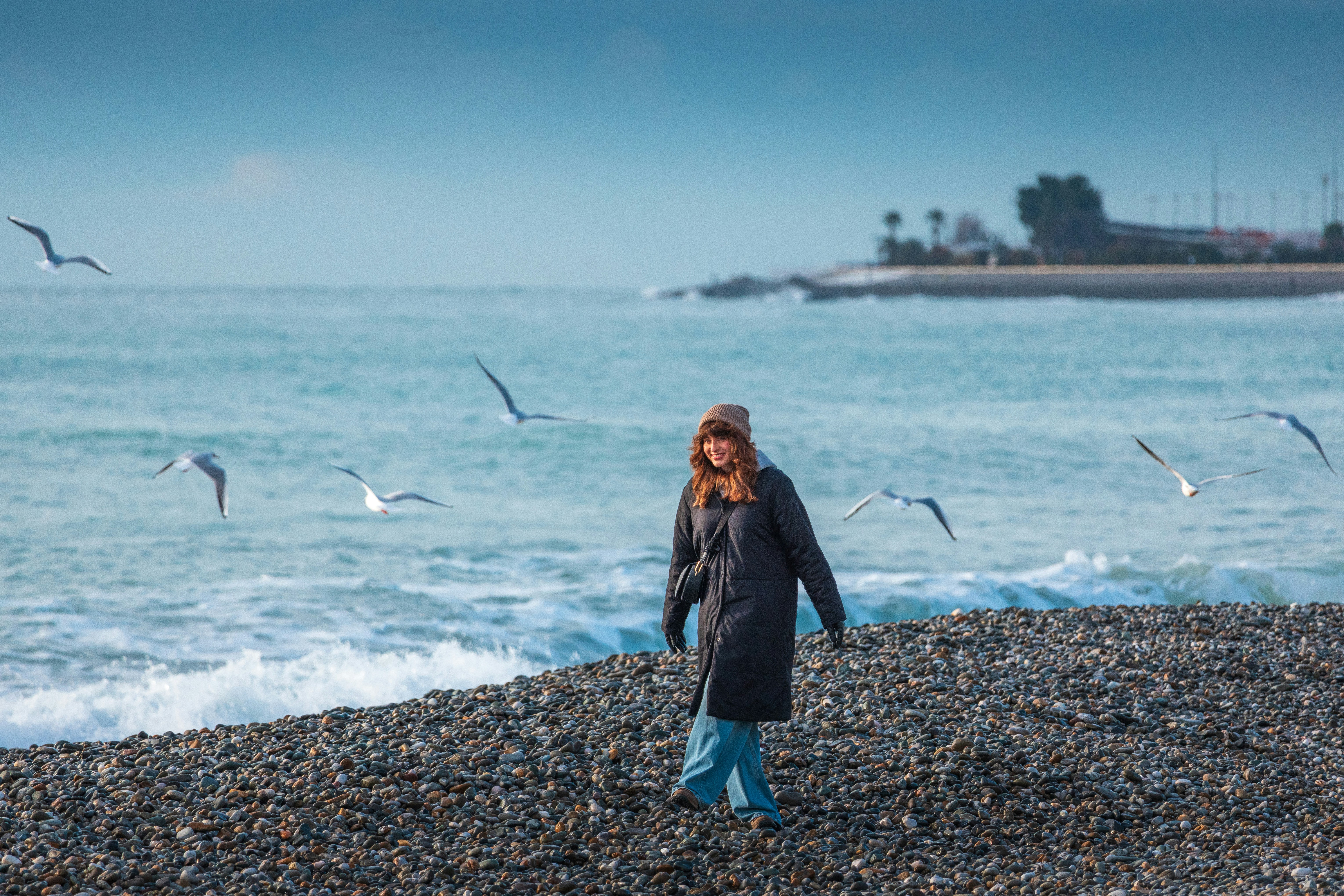 Woman walks on a pebble beach with seagulls flying.