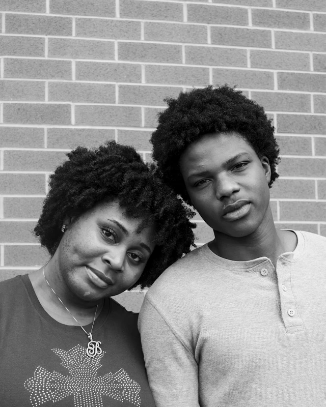 A mother and her teenage son leaning their heads together against a brick wall, in black and white