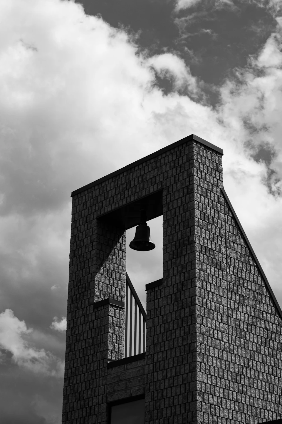 The image depicts a tall brick tower with a bell at its peak, situated against an overcast sky. The tower is constructed from large bricks and features a small window on one side. A black and white photograph captures the scene, emphasizing the contrast between light and shadow.