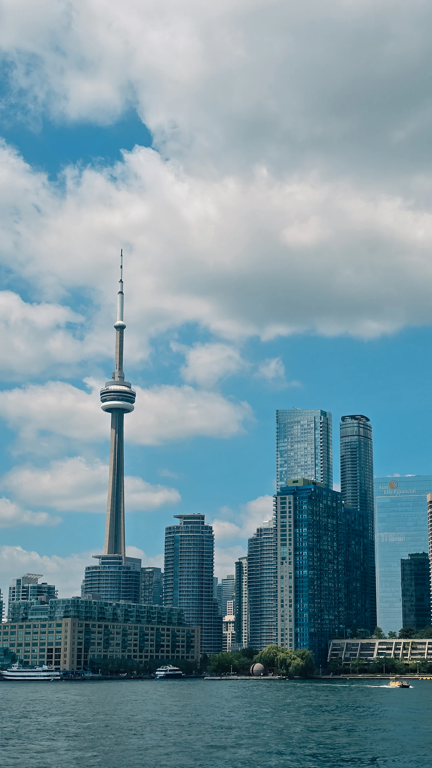 The Toronto skyline under a bright blue sky seen across the water, with the CN Tower and waterfront towers along Lake Ontario