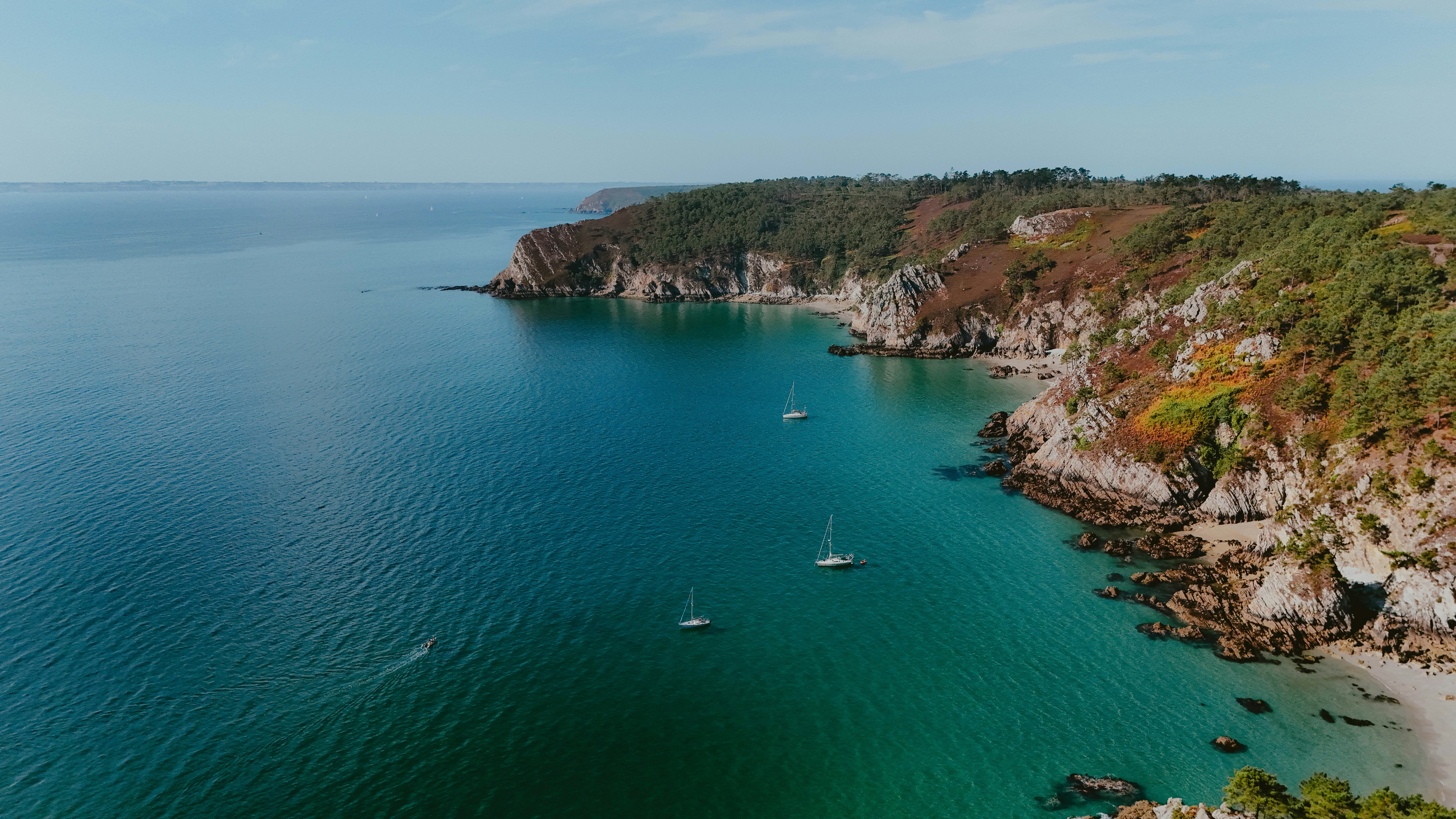 Sailboats glide on a turquoise bay near rocky cliffs.