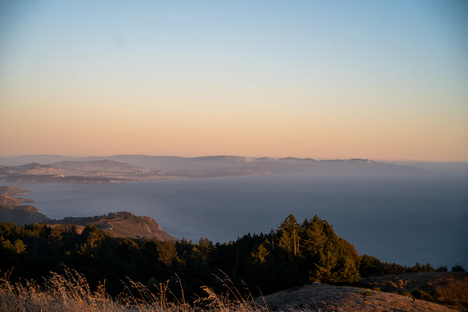The image depicts a serene landscape with a panoramic view of a coastal area, featuring a deep blue ocean and a light brown shoreline dotted with trees. The sky above is painted in hues of orange, suggesting either sunrise or sunset. The perspective from the top of a hill provides an expansive view of the surrounding terrain, showcasing the vastness of the landscape and the beauty of nature's elements coming together to create this picturesque scene.

The image does not contain any discernible text or countable objects, making it a purely visual representation of the natural environment.