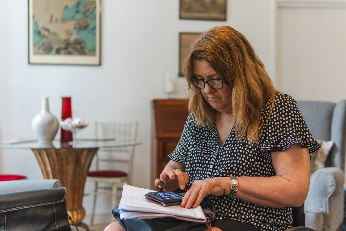 Une femme assise dans un salon étudie des documents posés sur ses genoux, une calculatrice à la main