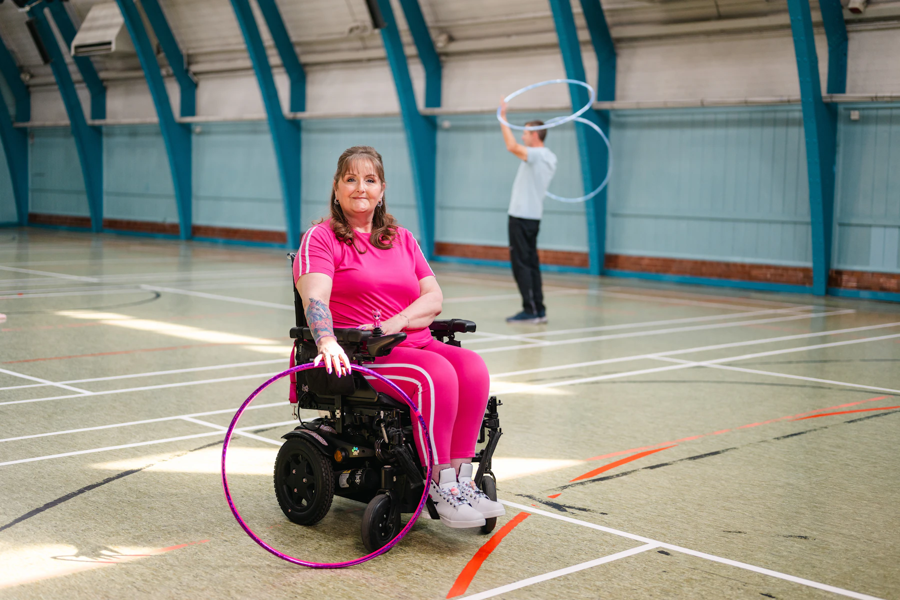 A woman in a powered wheelchair holding a pink hula hoop on the painted floor of a sports hall