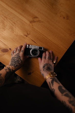 Tattooed hands hold a vintage camera on a wooden table.
