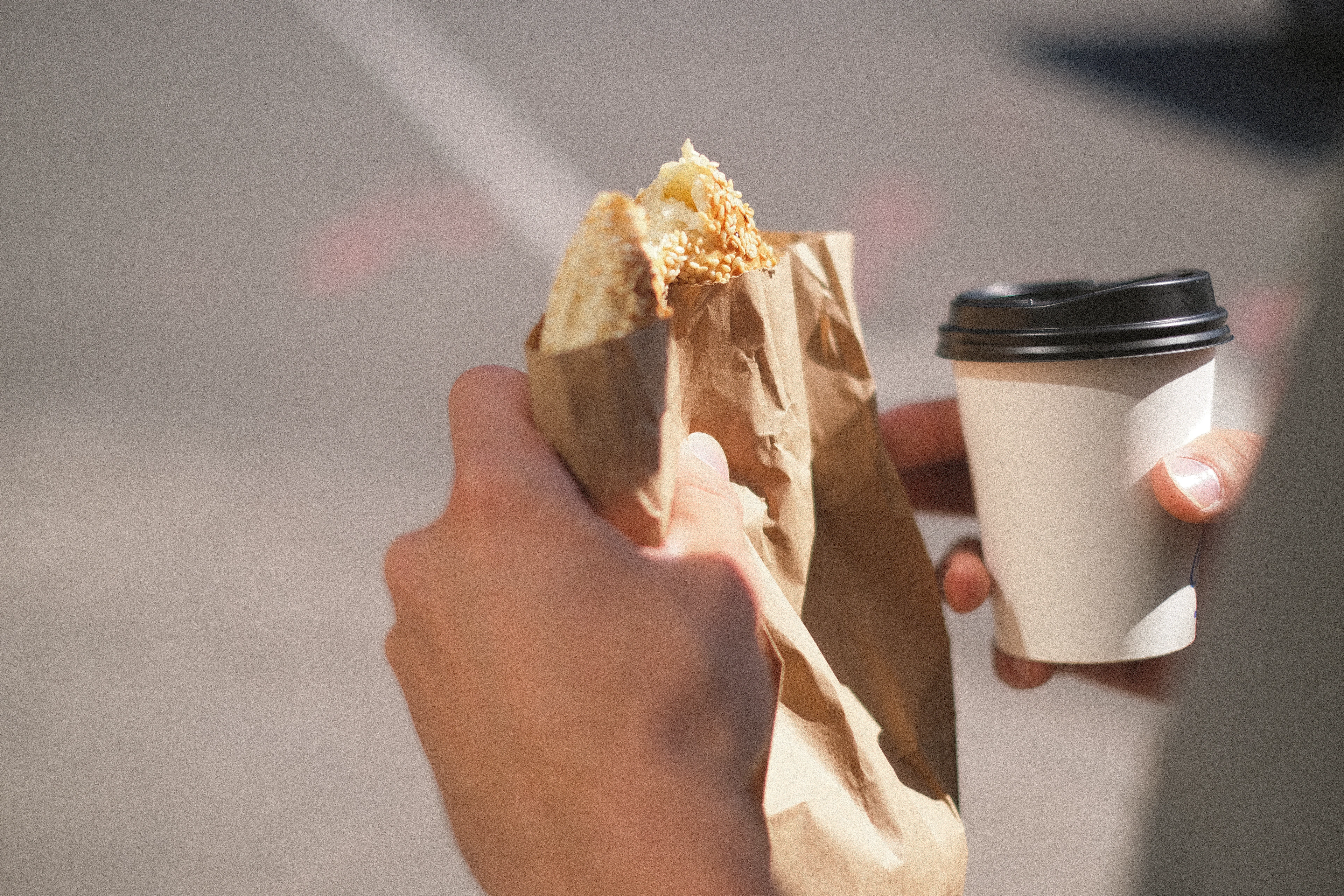 A hand-held Montreal bagel, hand-rolled and crusted with sesame seeds, half out of its brown paper bag, with a takeaway coffee cup in the other hand on the street.