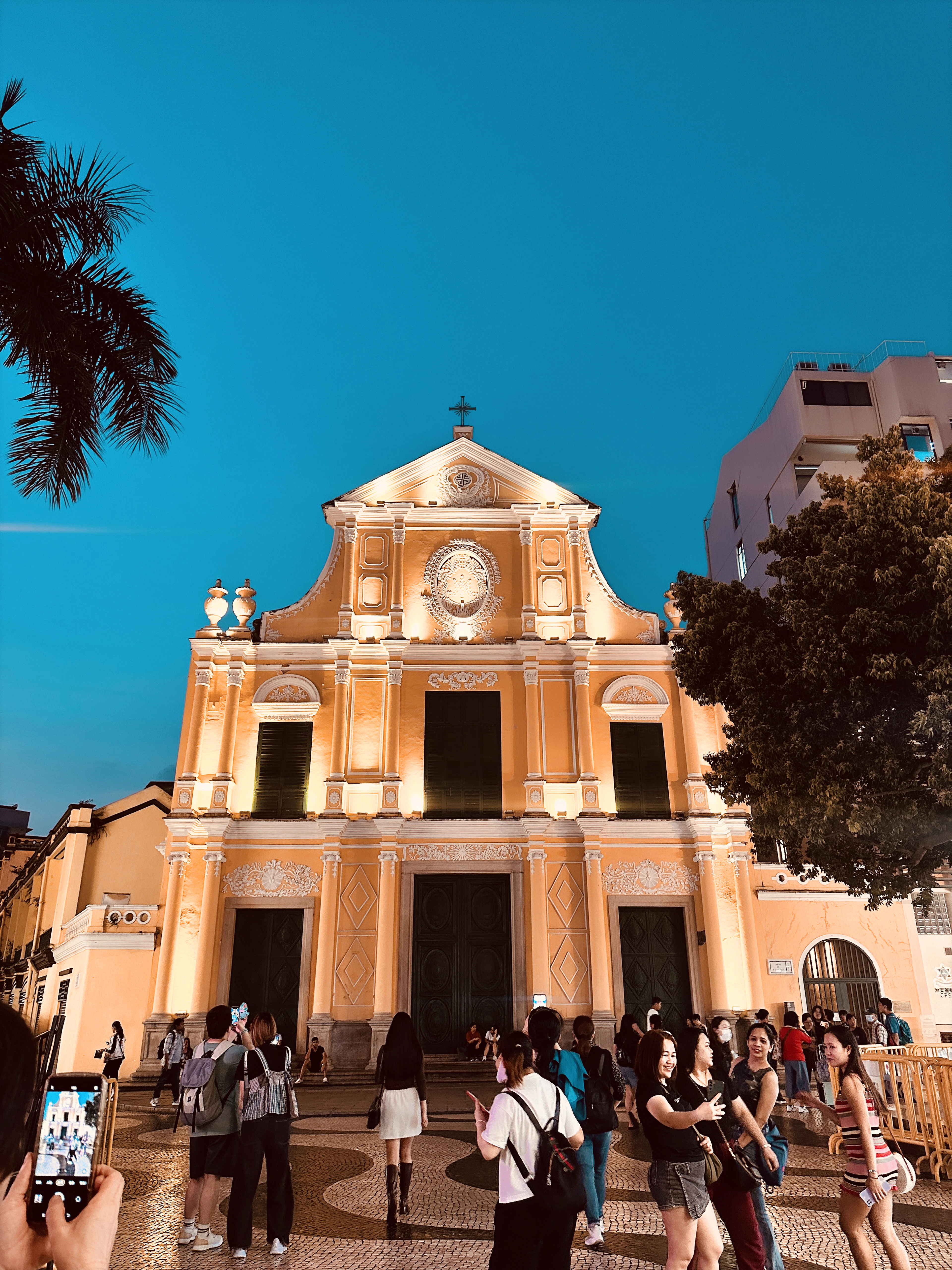 A mustard-orange baroque church facade lit at dusk against a deep blue sky, visitors gathered outside.