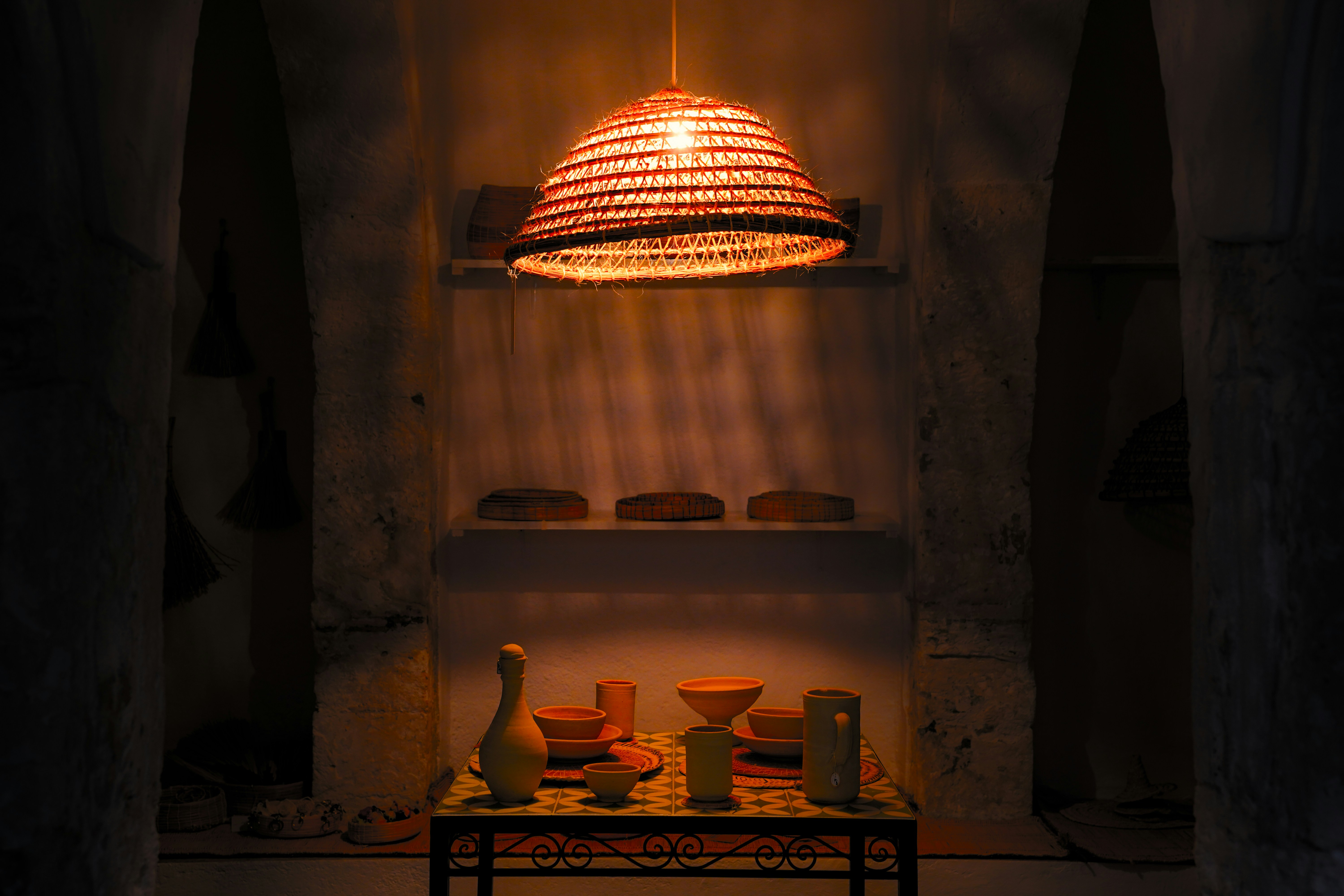 Woven lamp illuminates rustic display shelf with pottery.