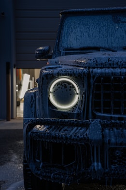 Dark suv covered in foam with glowing headlight.