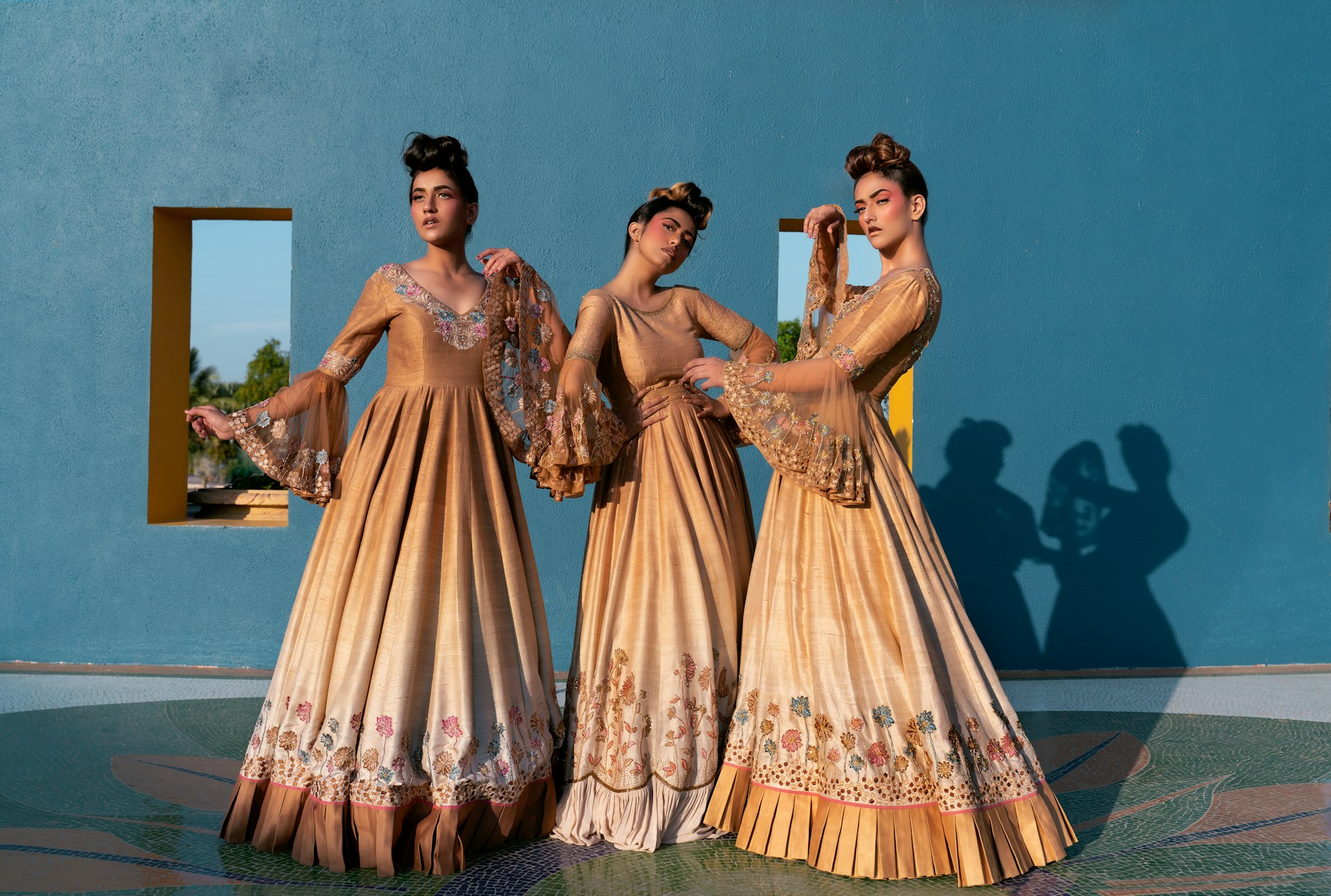Three women in golden gowns stand against blue wall.