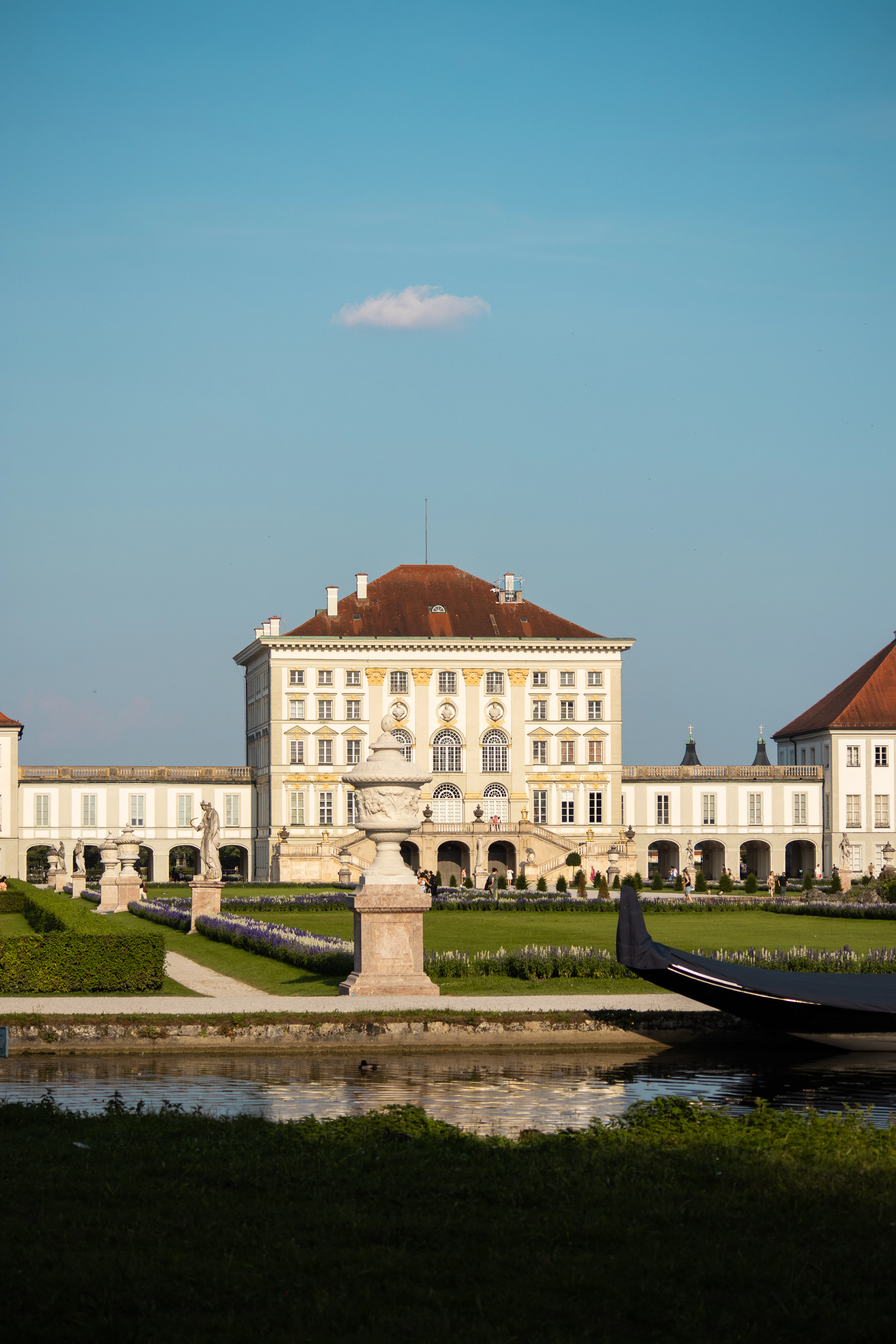 Nymphenburg Palace in Munich in low evening sun, its long baroque wings and statue-lined parterre mirrored in the still canal that runs up to the entrance