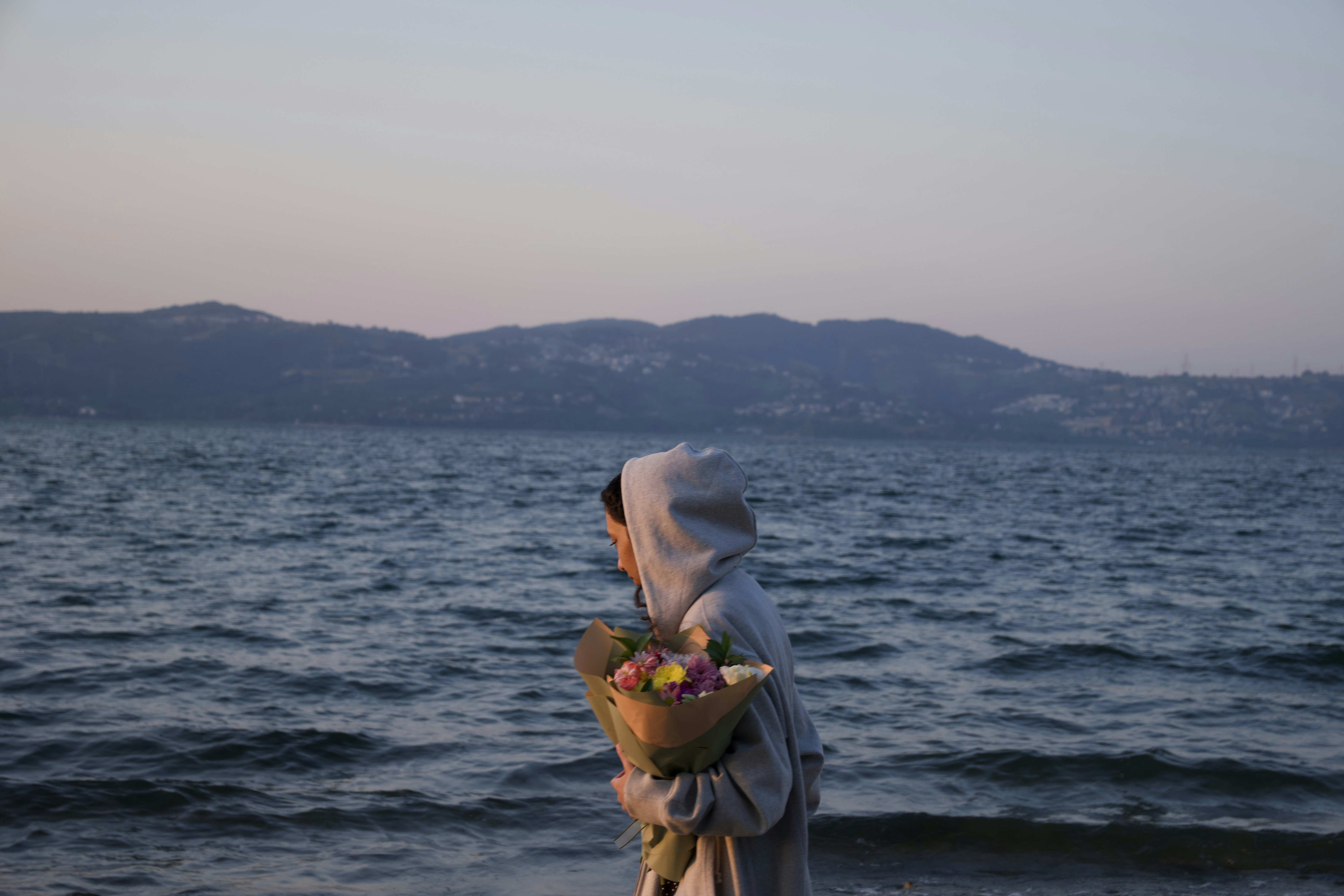 Person in hoodie holding flowers by the sea