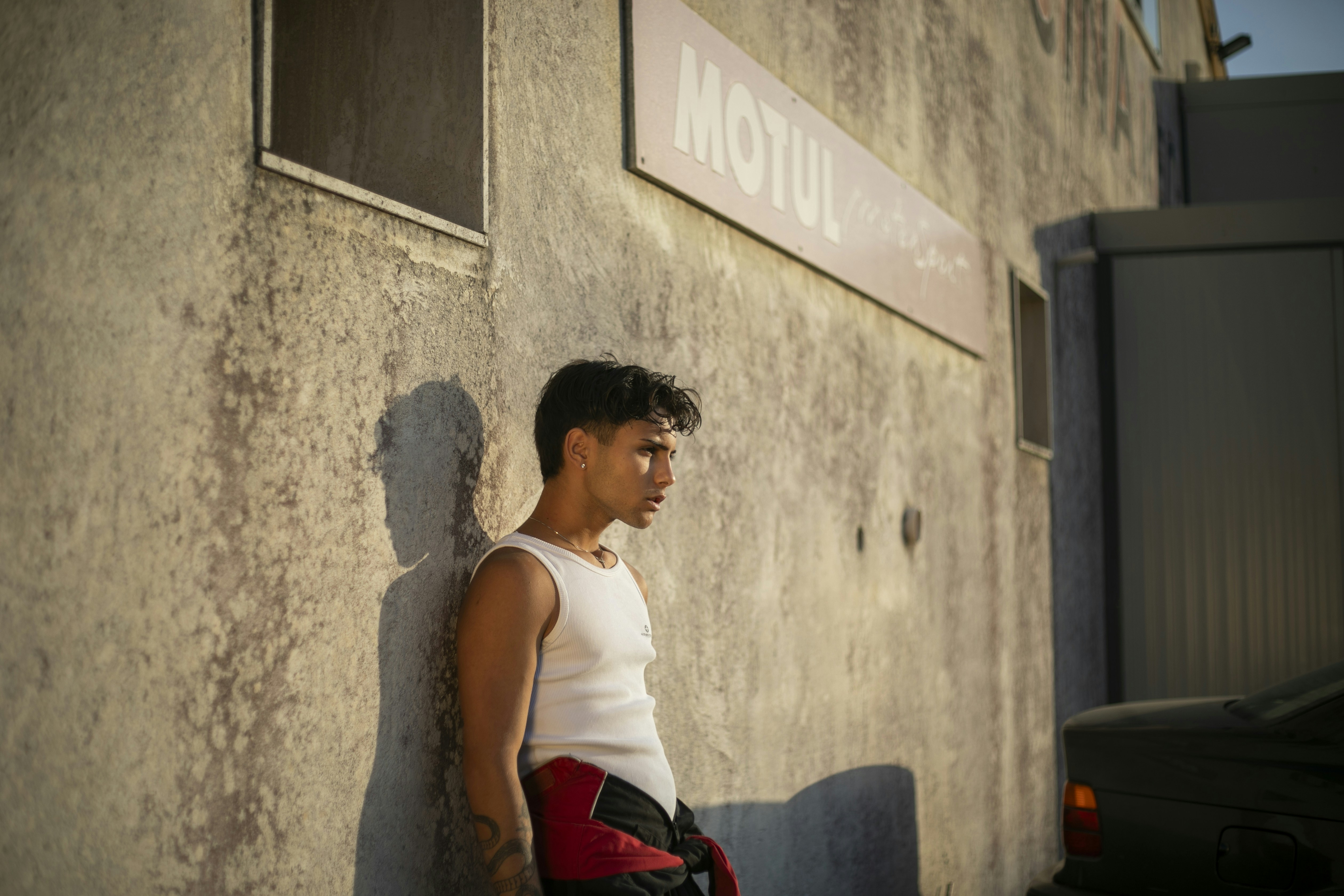 Man in tank top leans against a weathered wall.