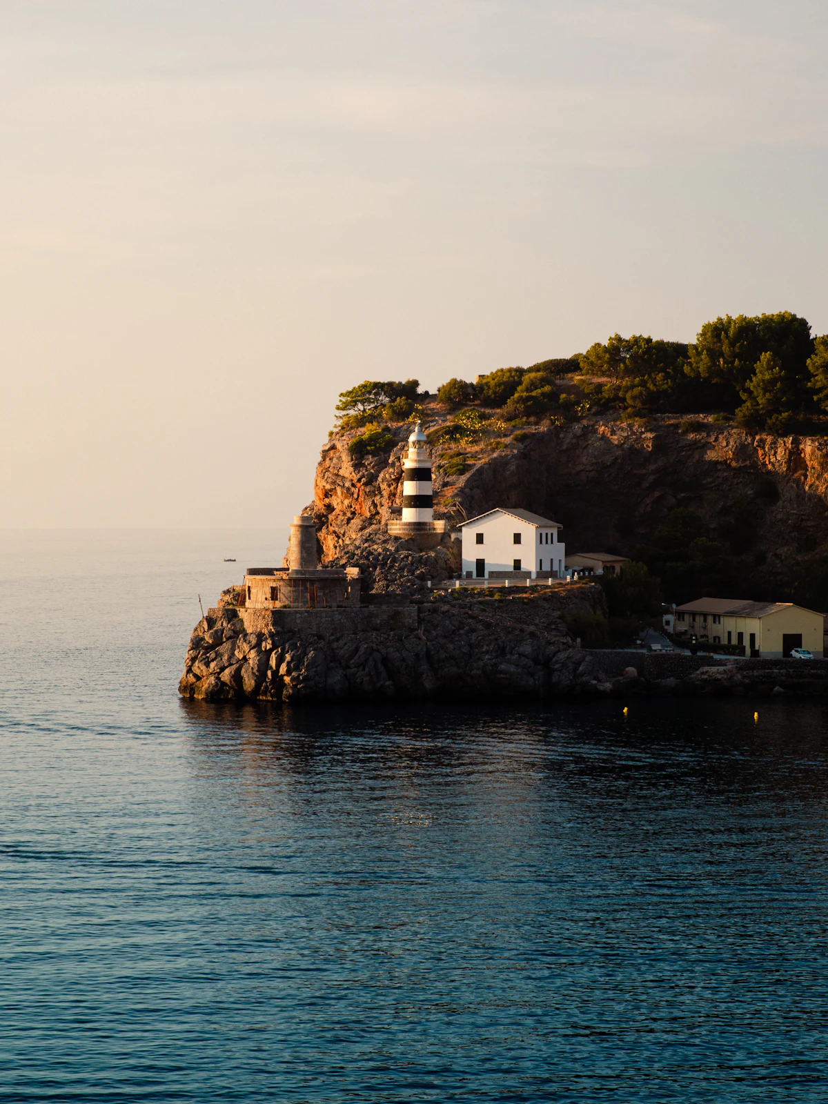 A striped lighthouse on a rocky point at Port de Sóller, with calm sea in low sun