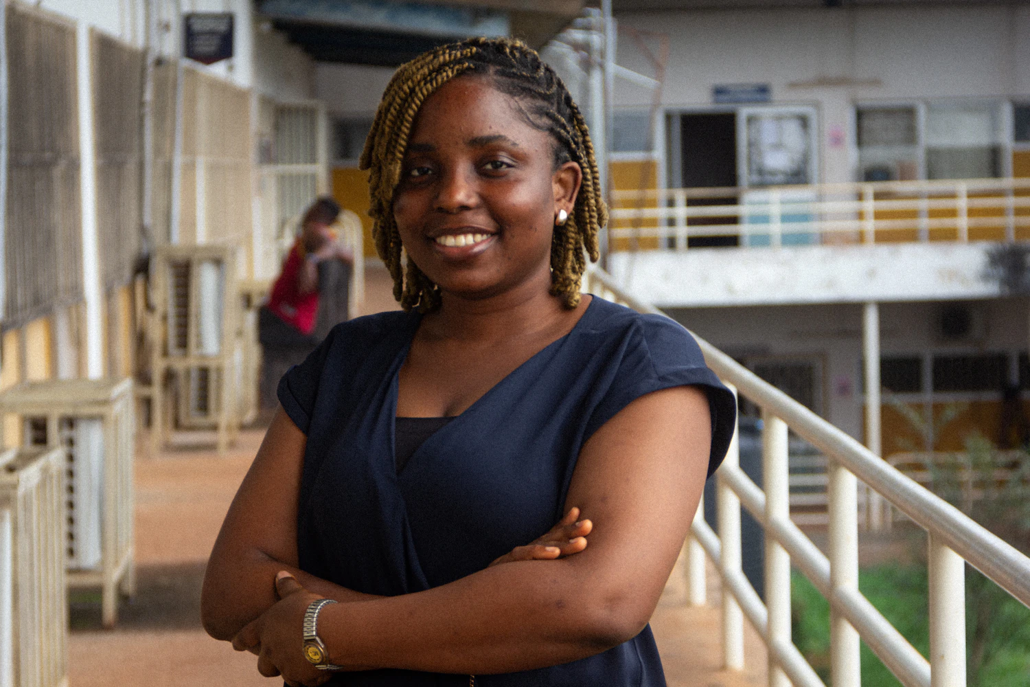 A woman with braided hair and folded arms standing on a steel walkway above a works floor