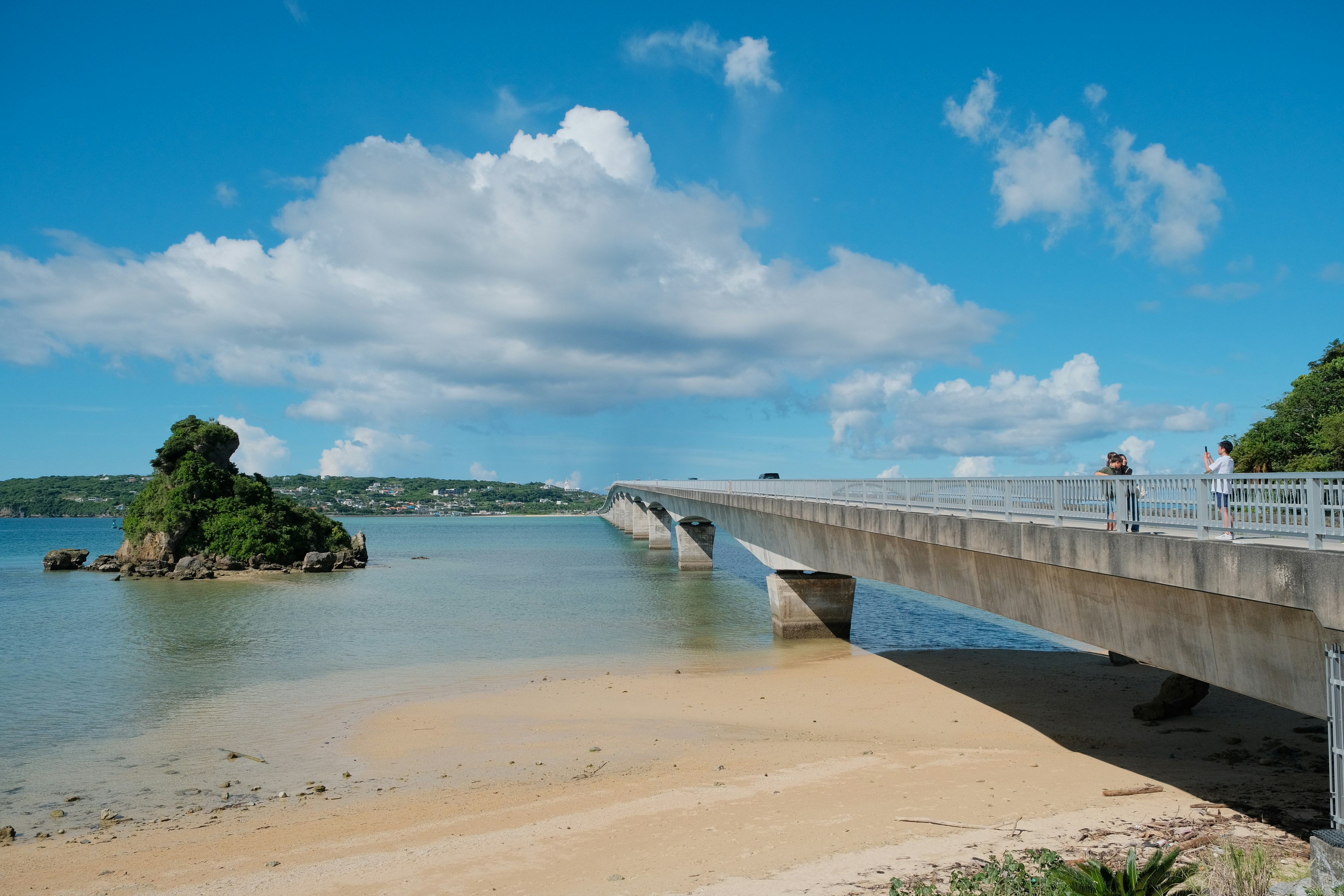 A concrete bridge crosses calm water near a sandy beach