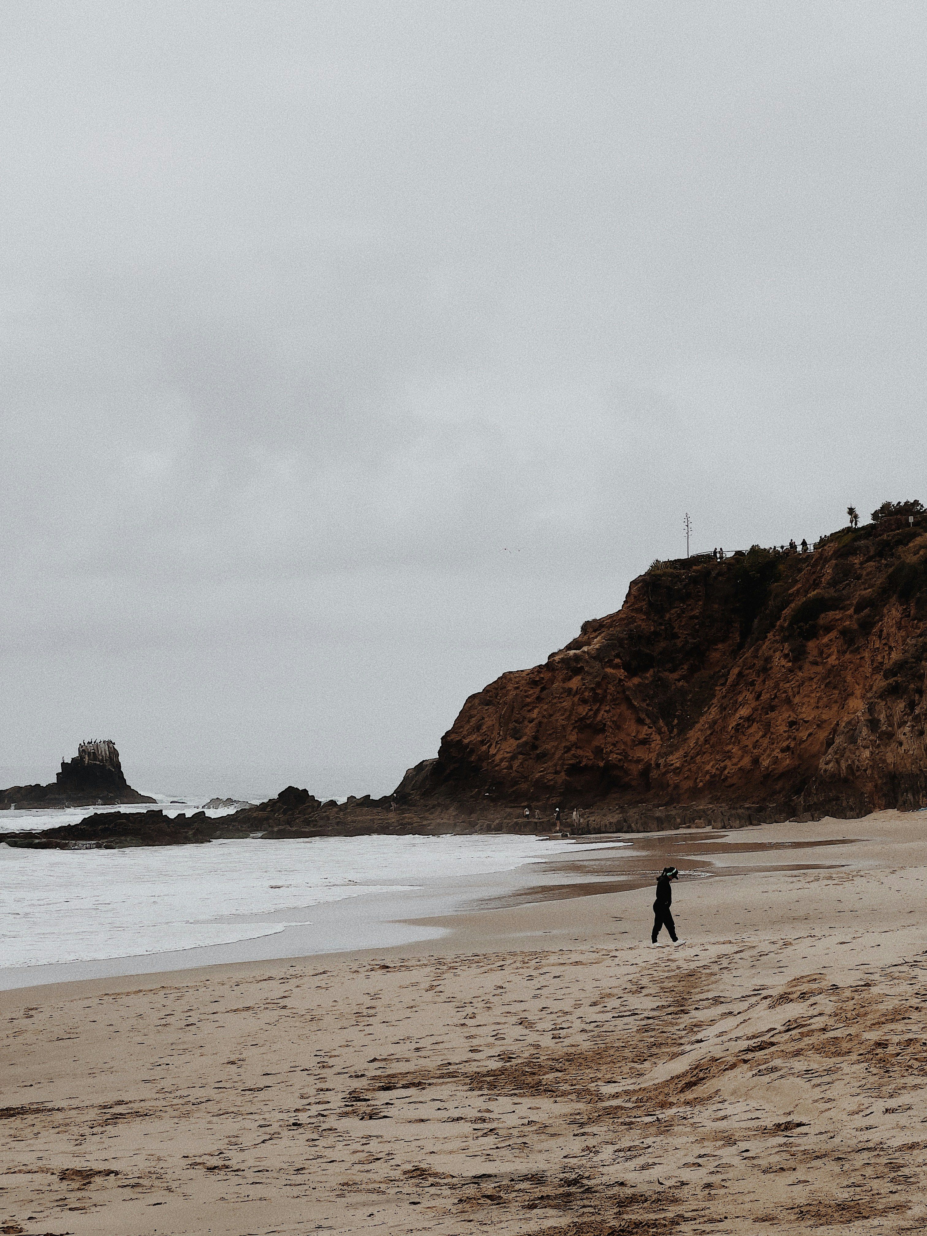 A lone figure walks on a sandy beach near rocky cliffs.