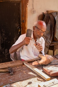 Elderly man carves wood with hammer and chisel.