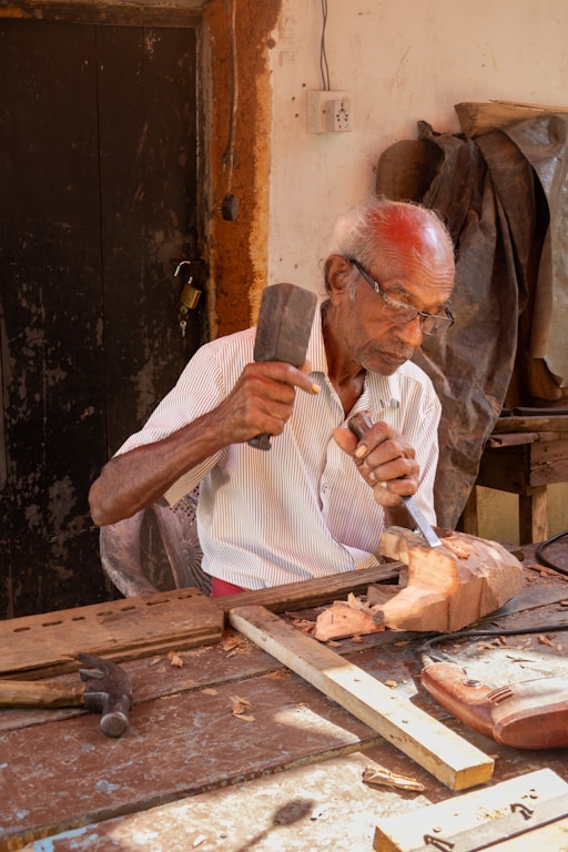 Elderly man carves wood with hammer and chisel.