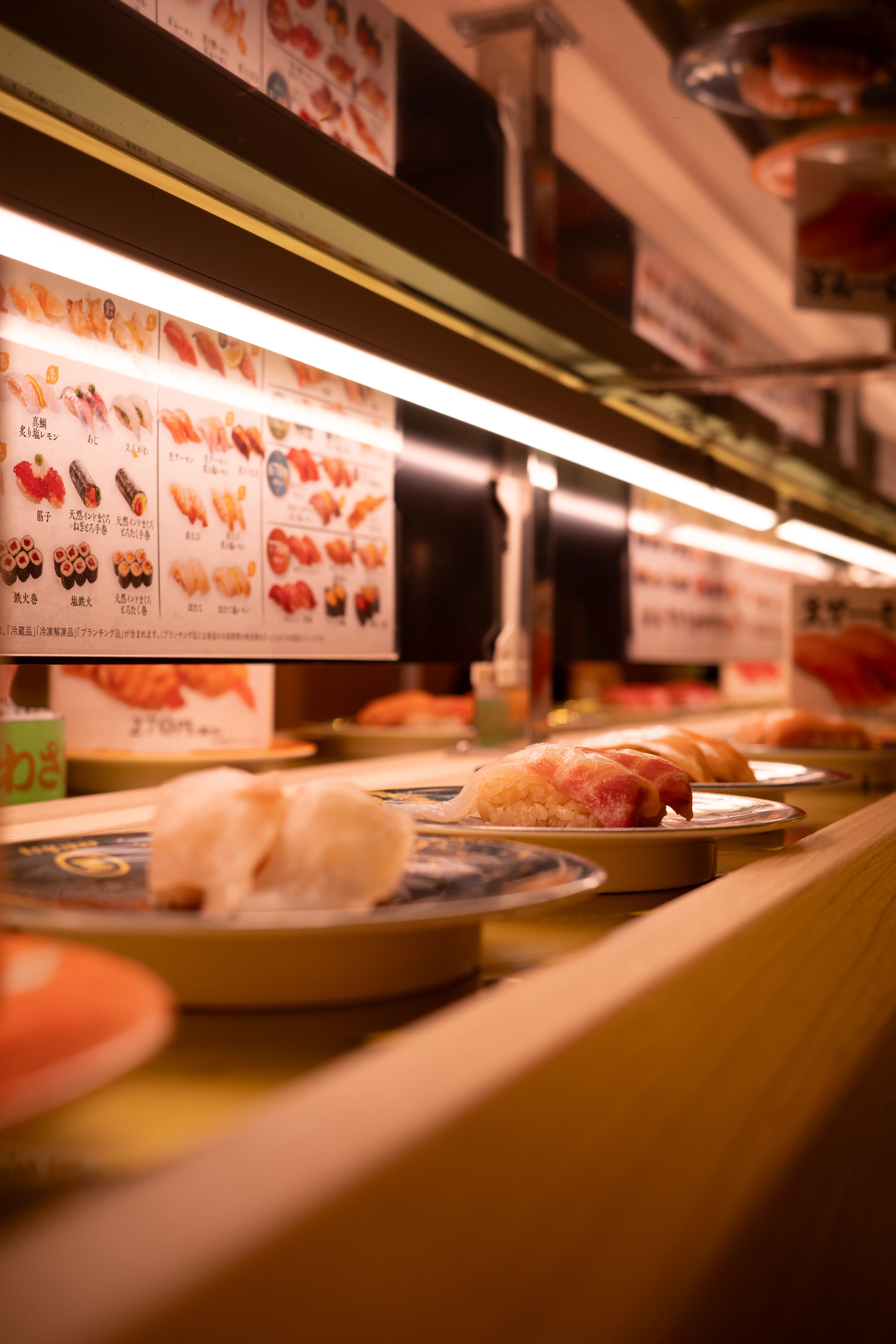 Plates of nigiri gliding along the conveyor belt of a kaiten sushi counter, lit Japanese menu boards with yen prices running above the track and a pale wood counter edge in the foreground