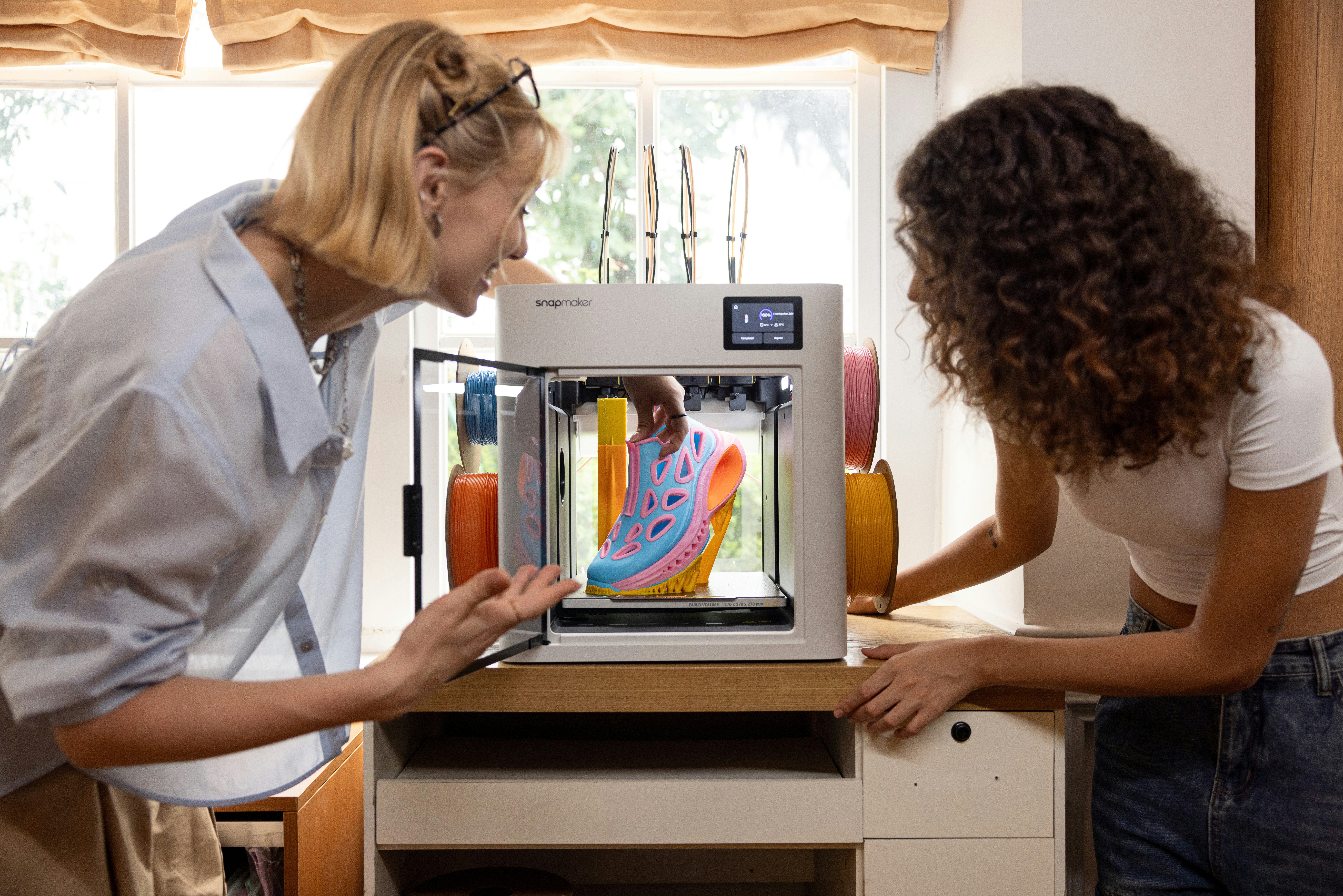 Two women observing a colorful shoe being 3D printed