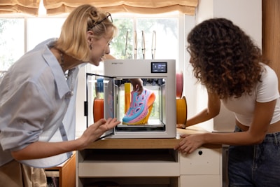Two women observing a colorful shoe being 3D printed