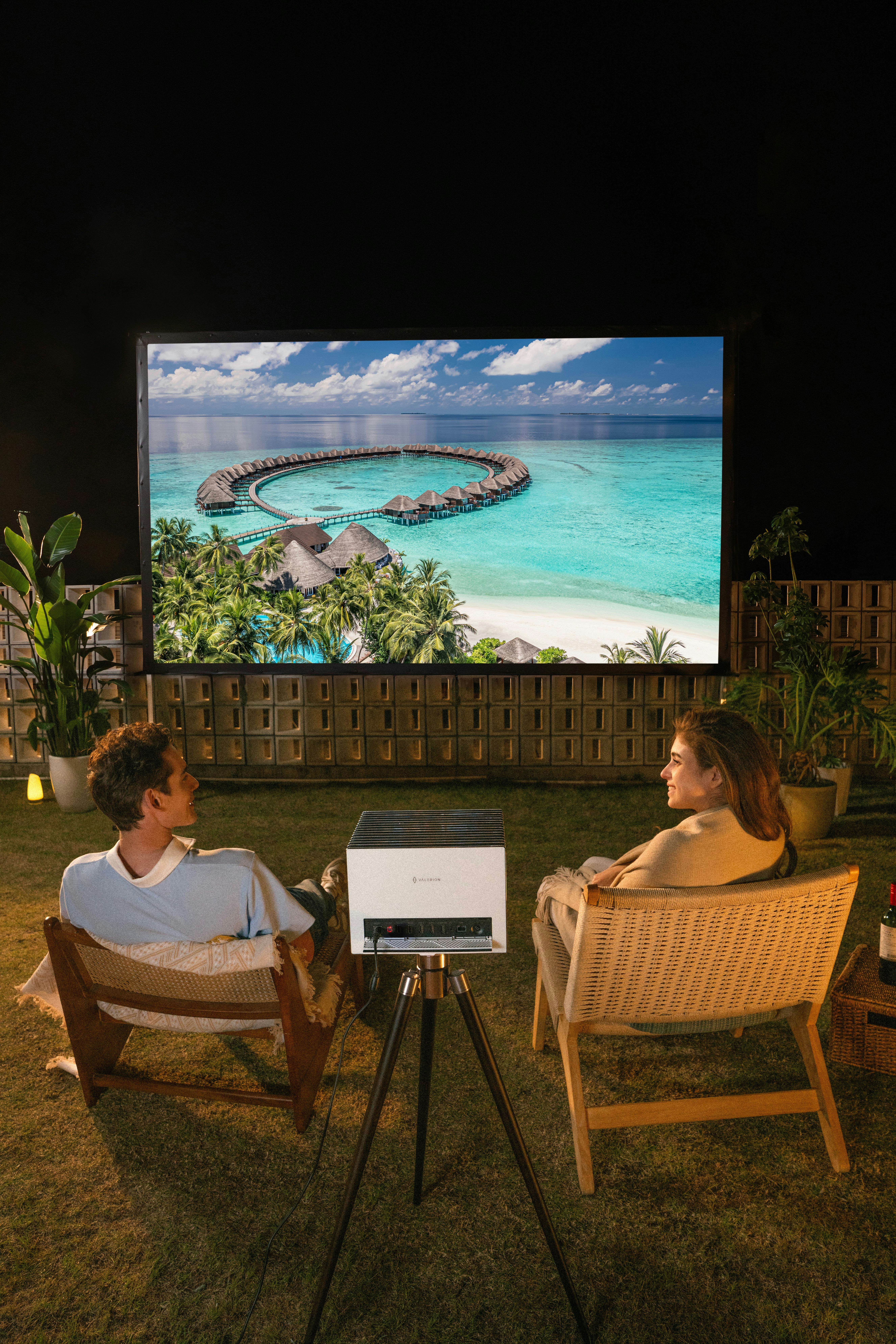 Couple watches movie on outdoor projector screen at night.