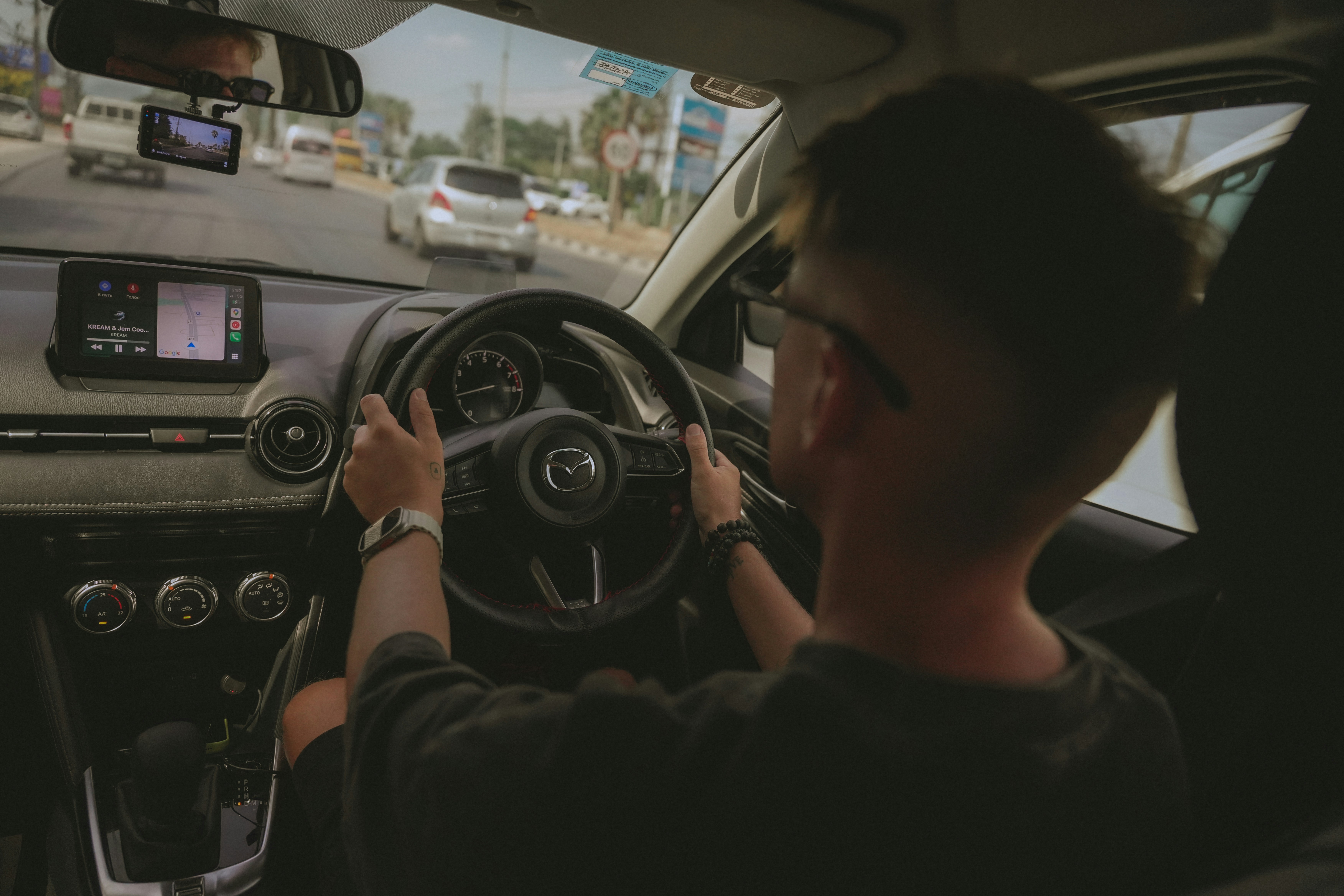 A man driving a car on a busy road, dashboard and steering wheel in focus with sunlight entering through the windshield.