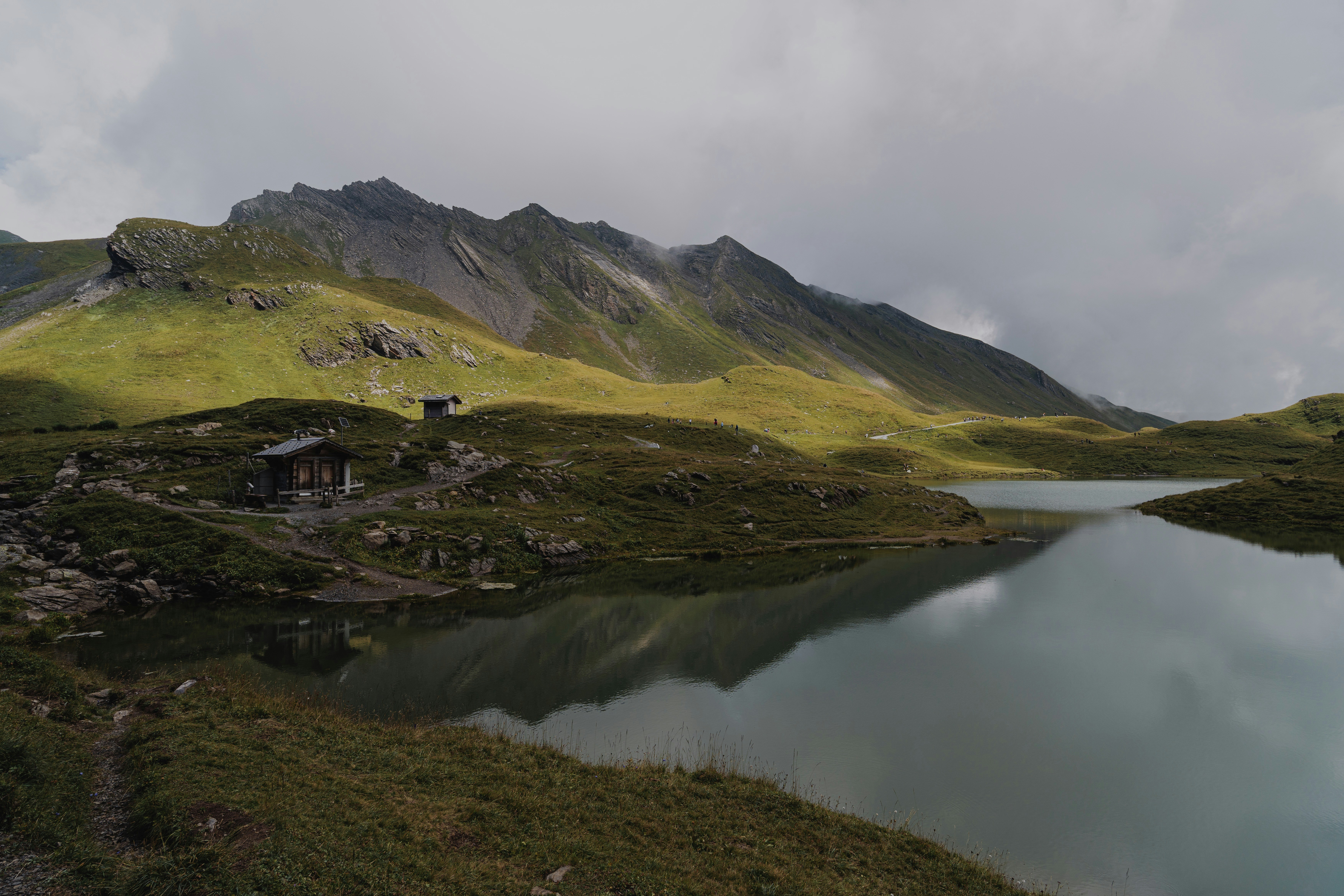 Wooden cabin by a lake with mountains under cloudy sky.