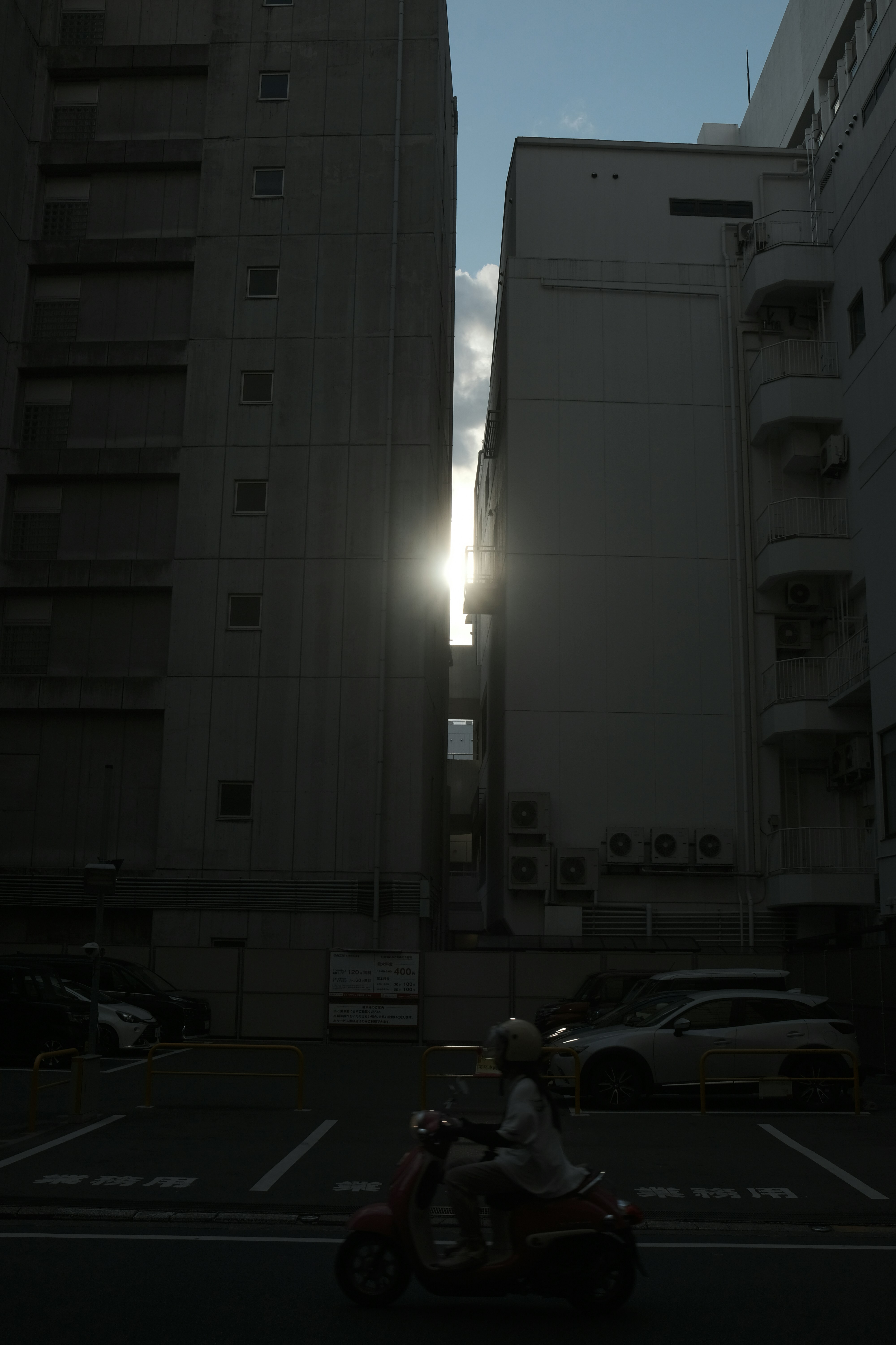 Motorcyclist passes between buildings with sun shining through.