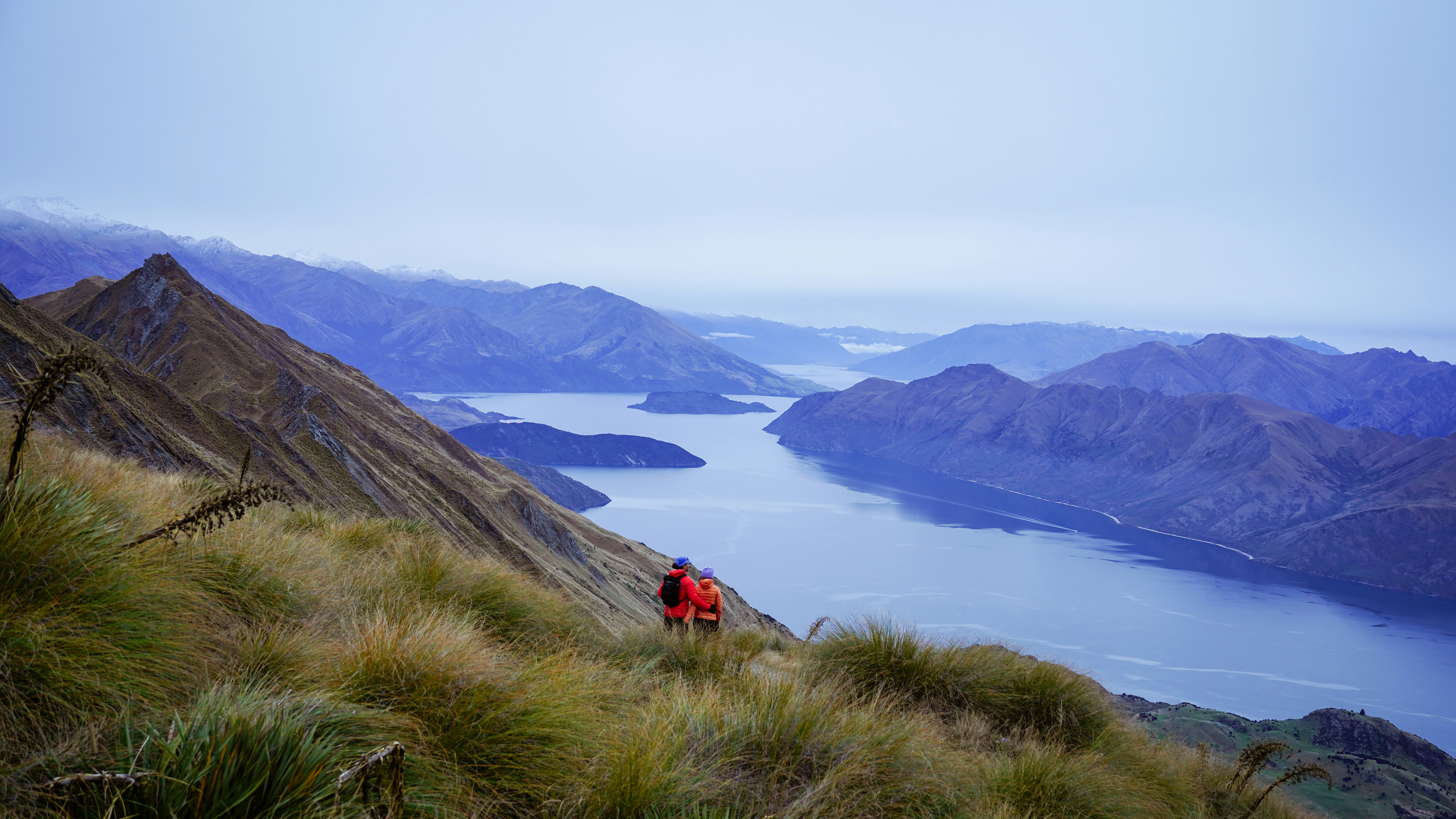 Two hikers ascend a grassy mountain overlooking a lake.