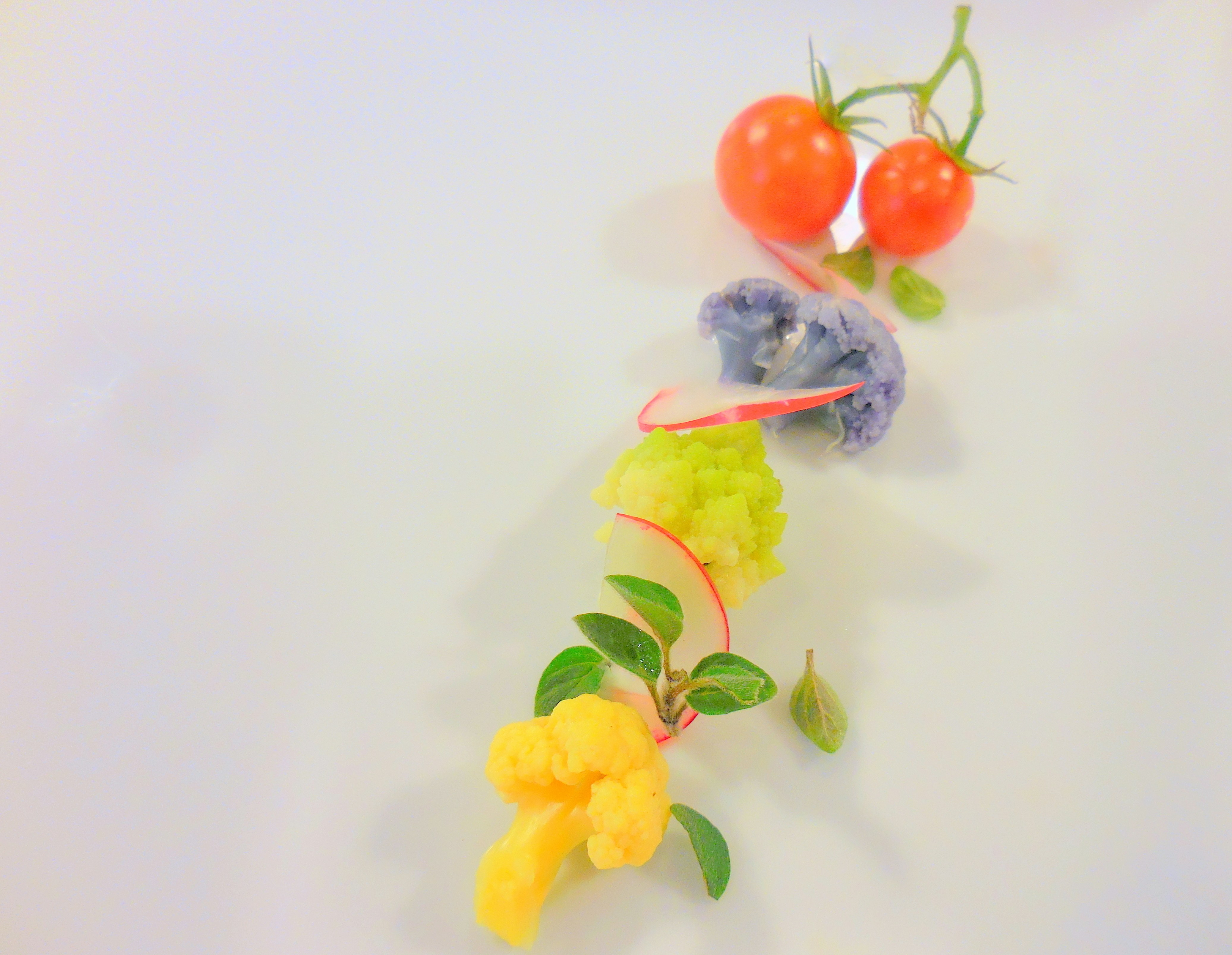 Colorful diagonal arrangement of cherry tomatoes, purple and yellow-green florets, and sprigs of leaves on a white background. Still-life photograph emphasizing color and texture.