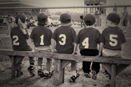 Five children in baseball uniforms sit on a bench.