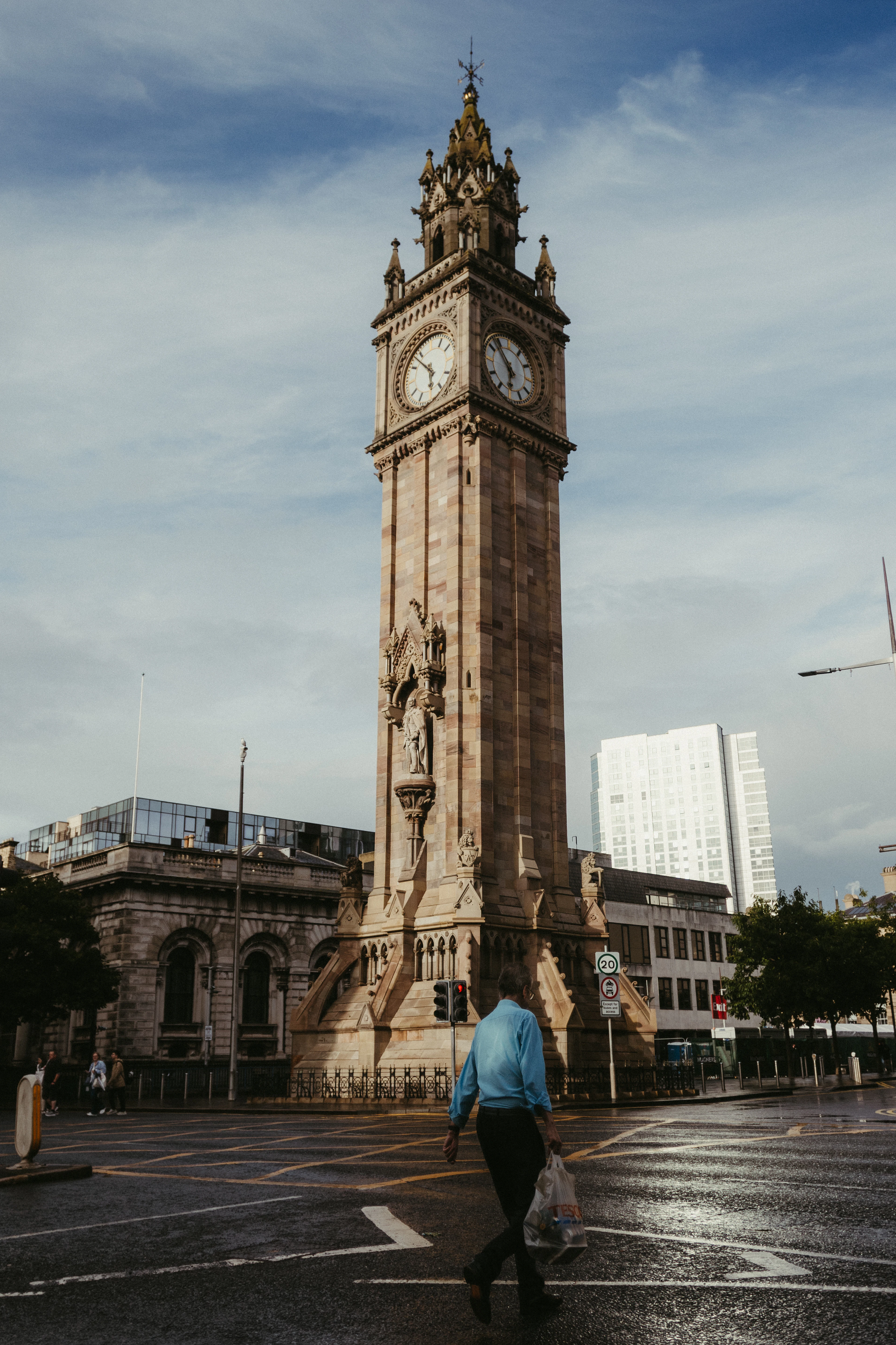 The Albert Memorial Clock tower in Belfast's Queen's Square lit warm against a blue sky, a man in a blue shirt crossing the rain-slicked road in front of it with a carrier bag.