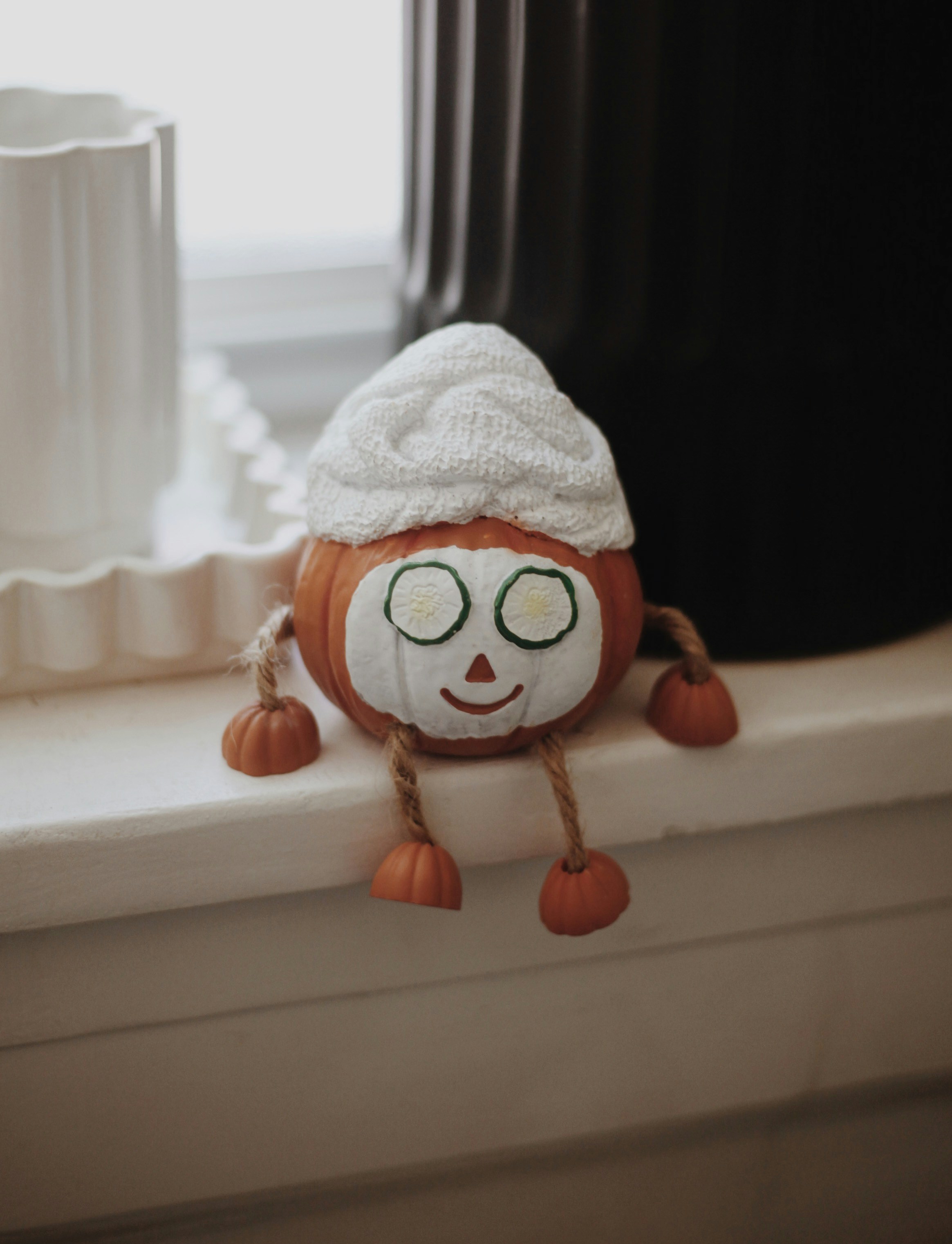 A cute pumpkin doll with a knitted hat sits on a windowsill.