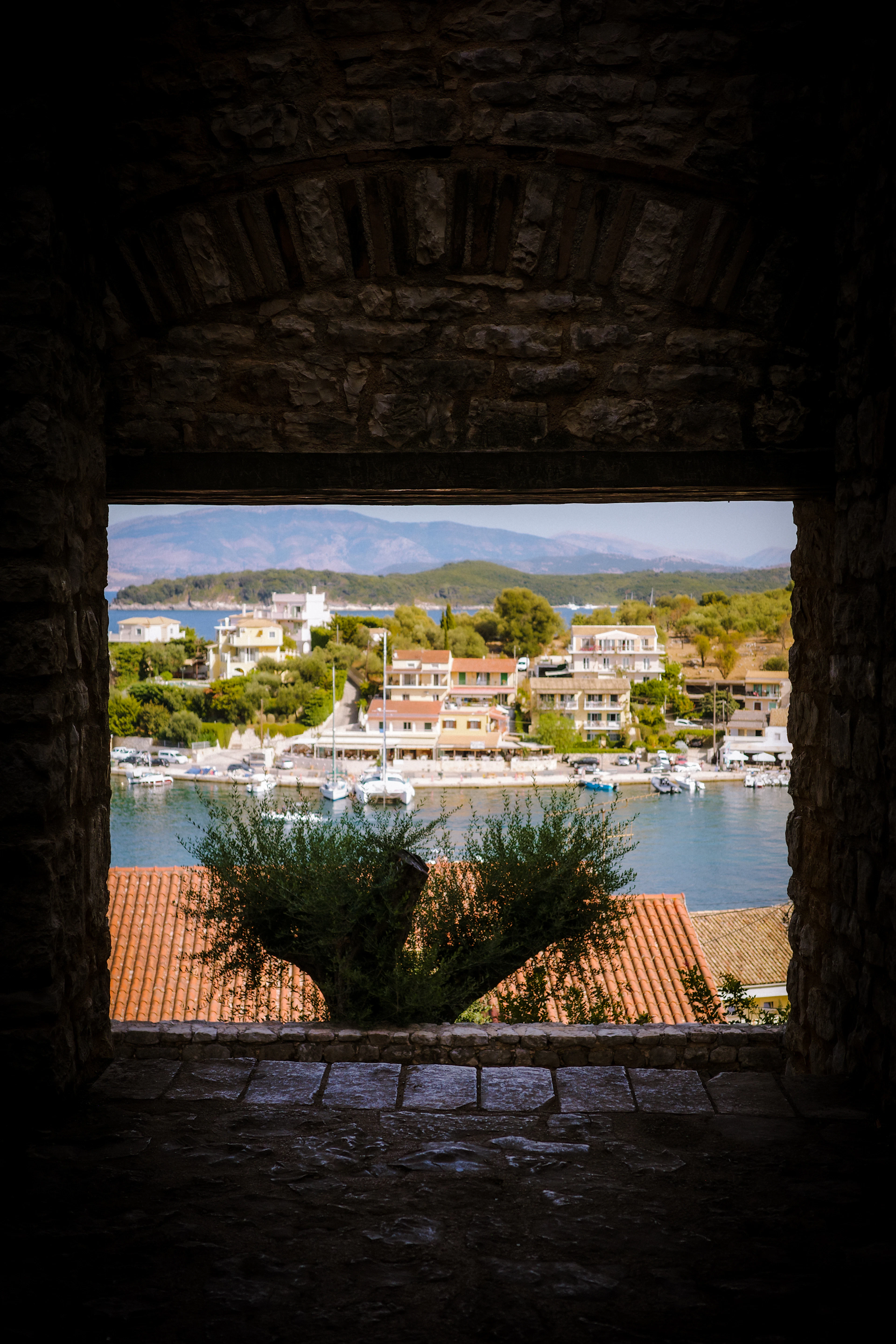 Kassiopi harbour on Corfu's northeast coast framed through a stone arch of the old castle, yachts moored along the quay and the mountains of the mainland hazy across the strait.