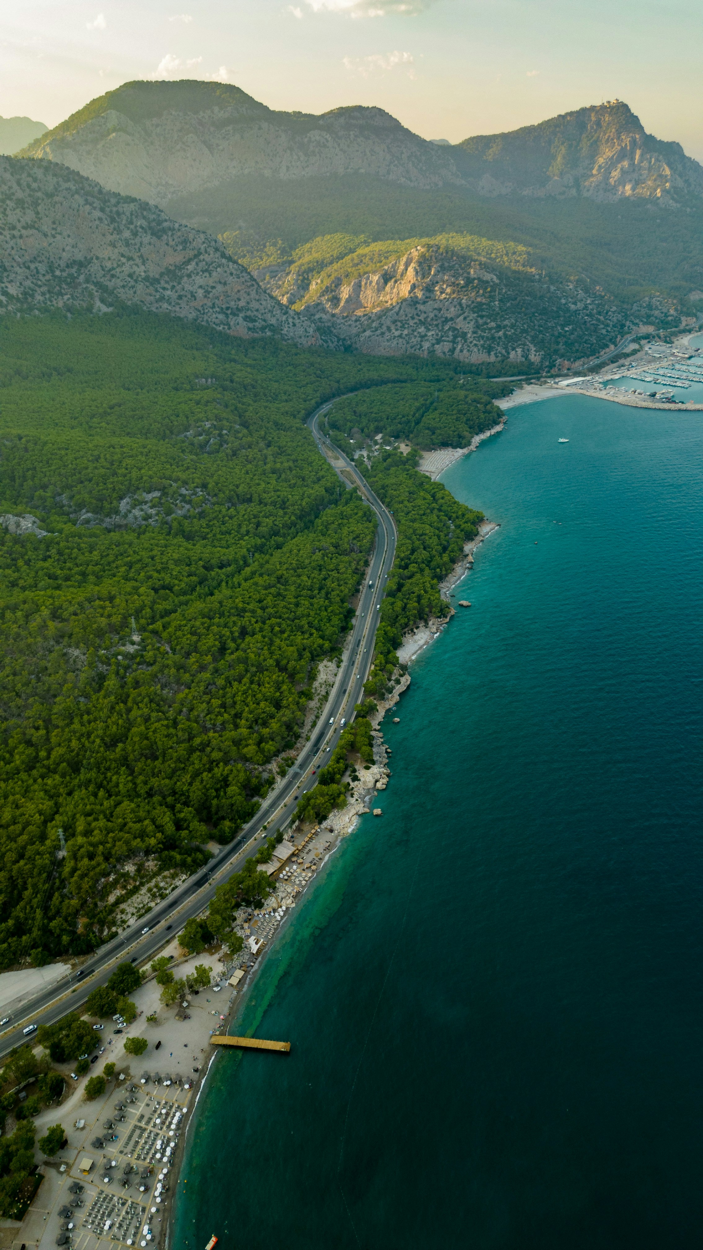 Coastal road winds through forest by turquoise sea and mountains.
