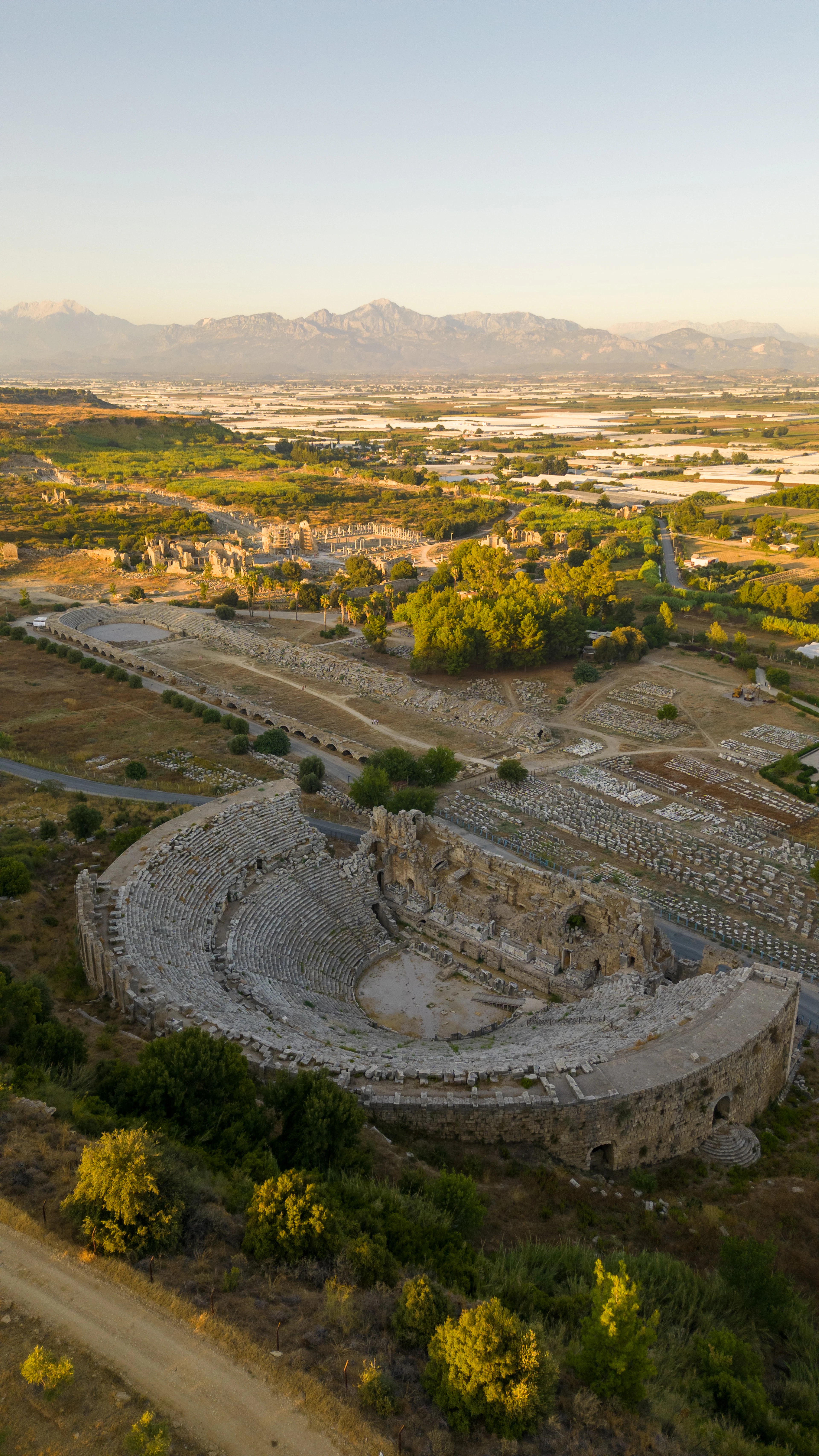 Aerial view of the ruined Roman theatre at Perge at golden hour, with the Taurus Mountains behind.