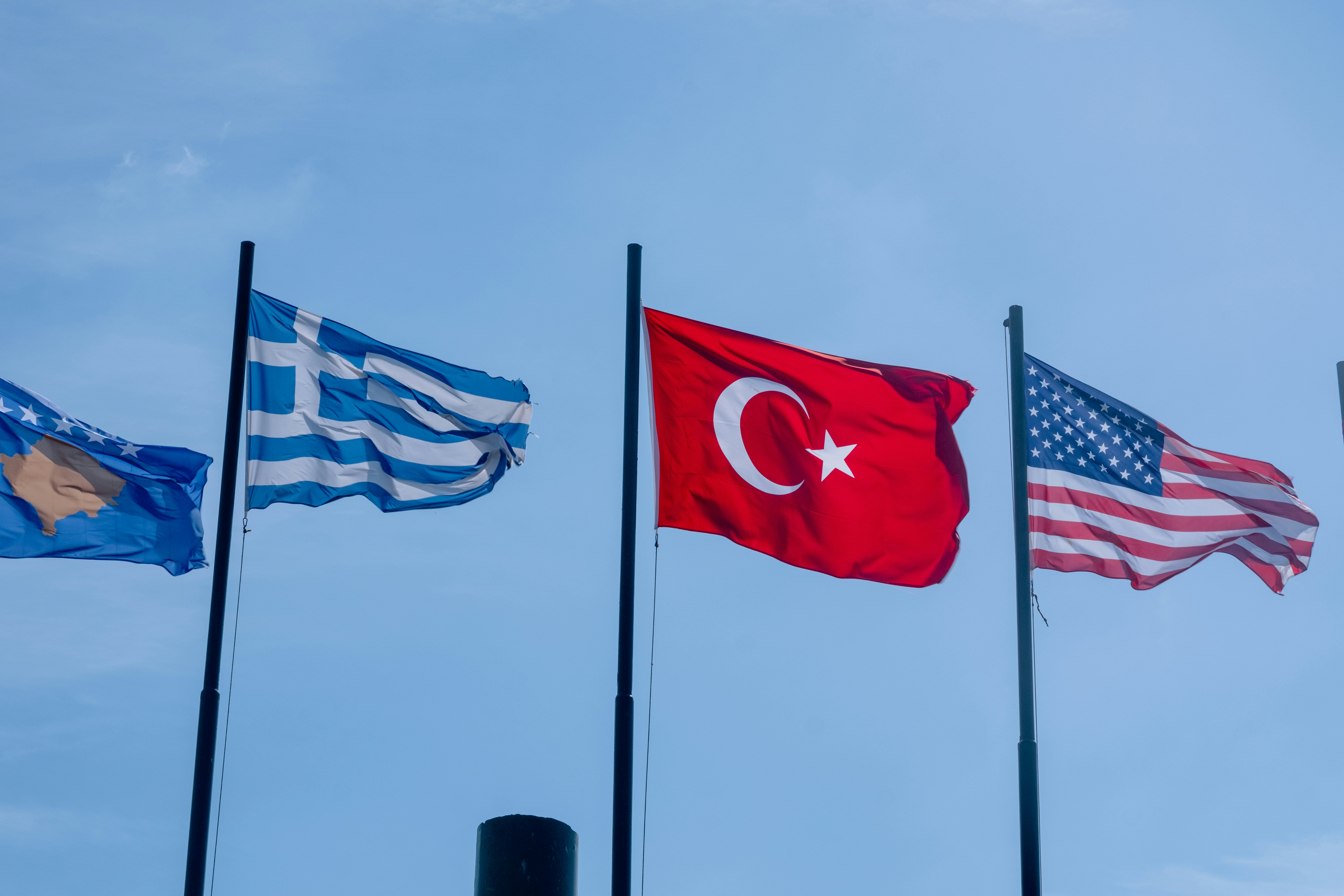 The flags of the European Union, Greece, Turkey, and the United States fly side-by-side against a partly cloudy sky.