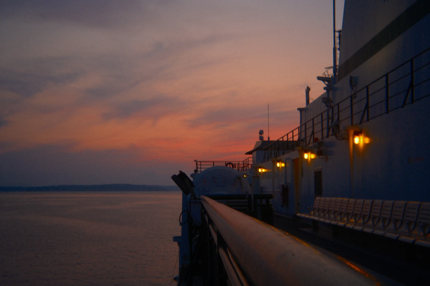 The deck of a ferry at dusk, cabin lights on above a pink sea