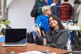 A smiling woman holds up two fingers near a laptop.